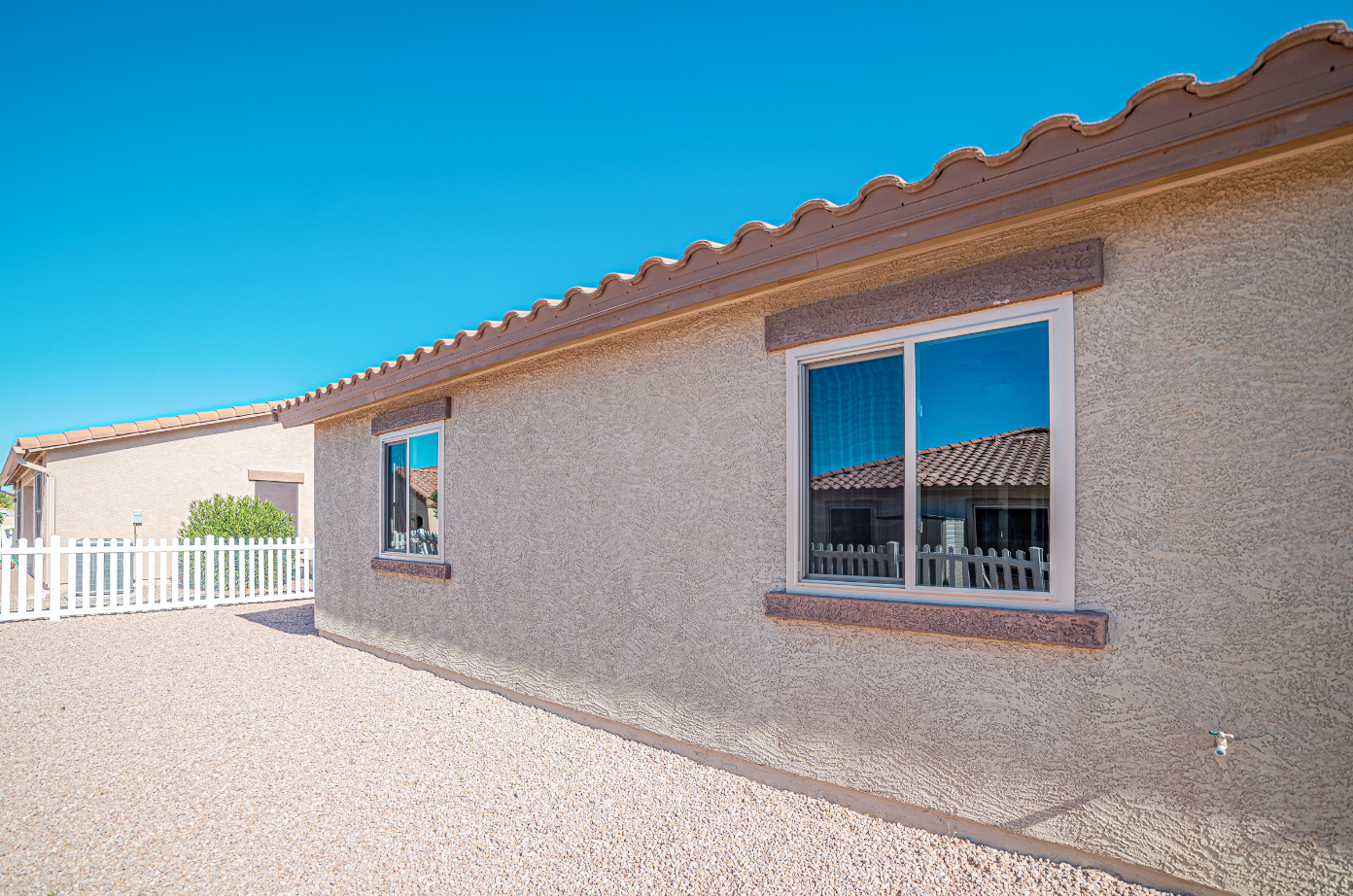 Exterior view of a beige stucco house with two windows reflecting blue sky and neighboring house, gravel ground, white picket fence, and a clear blue sky.