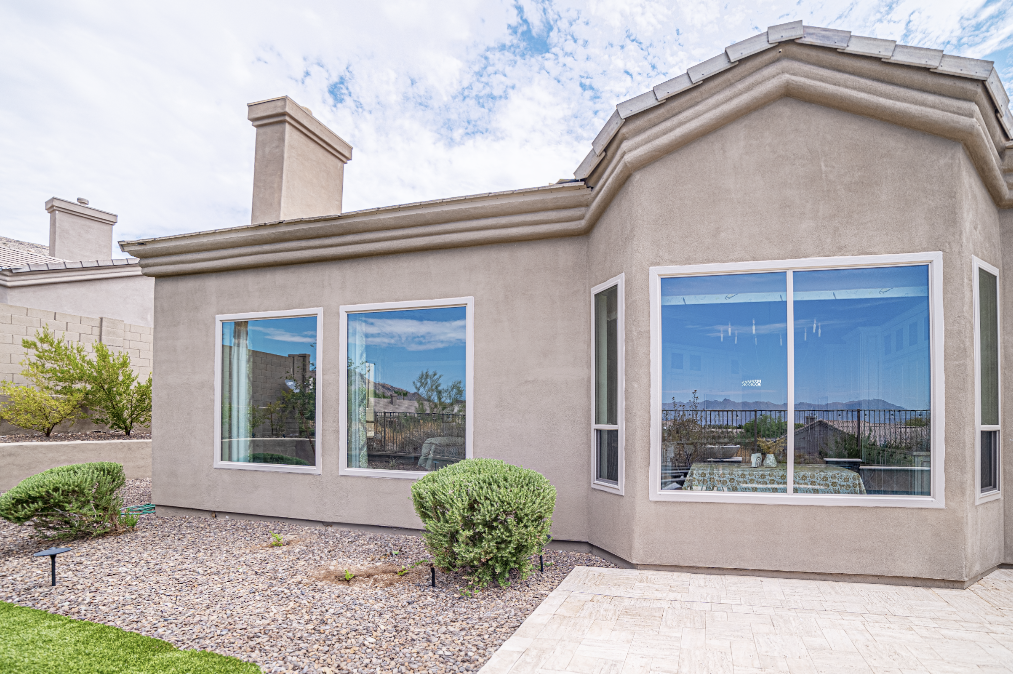 Backyard of a house with large windows reflecting the sky, mountains, and nearby houses, with desert landscaping and bushes.