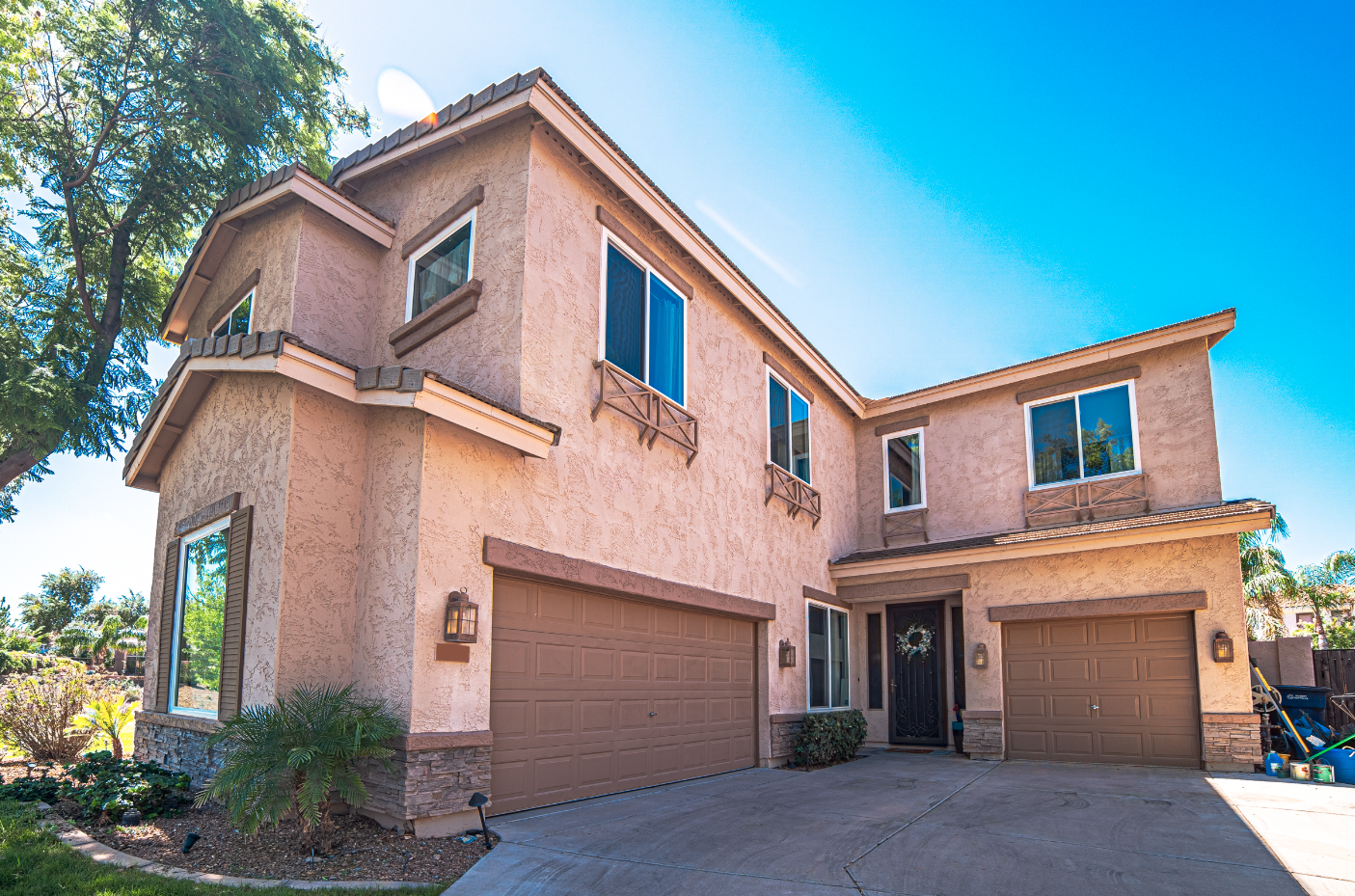 A beige multi-story house with a tiled roof, two garage doors, and a front door with a wreath, set against a bright blue sky with some trees in the background.