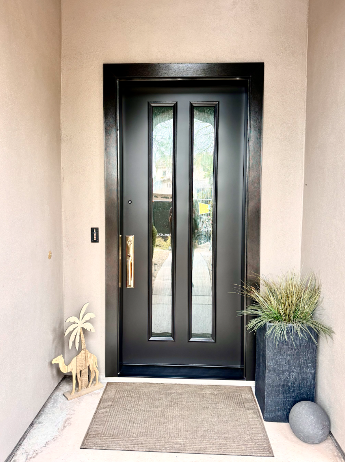 Black front door with glass panels, surrounded by beige walls, with a beige doormat in front, a decorative wooden camel and palm tree on the left, and a tall planter with greenery and a gray spherical ornament on the right.