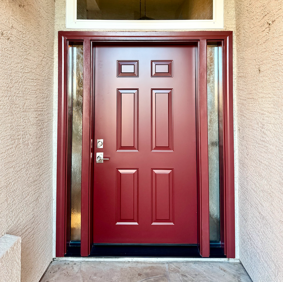 Red front door with decorative rectangular panels, chrome handle and lock, flanked by glass sidelight panels, with beige stucco walls and a concrete porch.