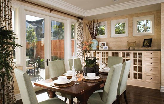 Dining room with a wooden table set for four, surrounded by light-colored chairs, large sliding glass doors leading to a patio, and small windows with a view of trees outside.