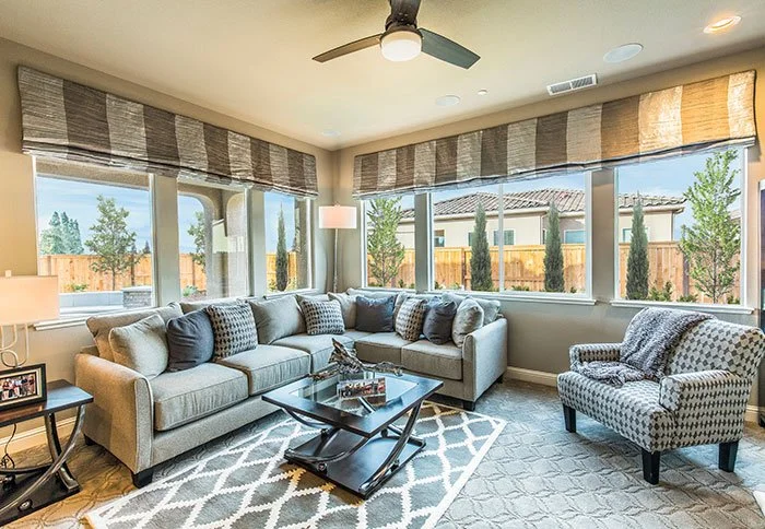 Bright living room with large windows, a gray sectional sofa with pillows, a patterned armchair, a black glass coffee table, a side table with a lamp, and a ceiling fan.