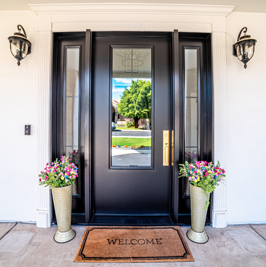 Front entrance of a house with a black door, two tall flower pots with pink and purple flowers, a welcome mat, and wall-mounted lanterns.