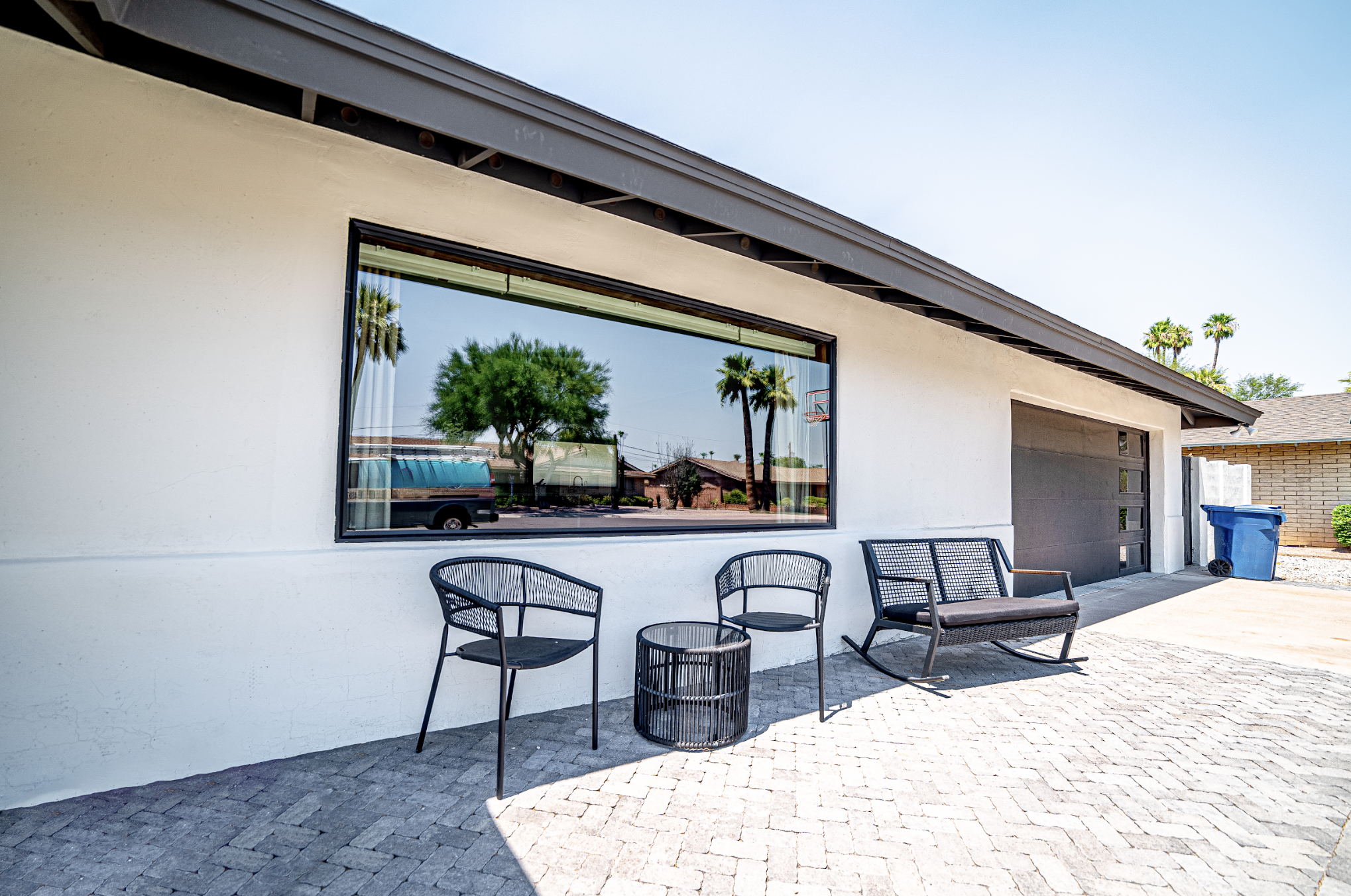 Modern house patio with black outdoor chairs, a small round table, and a black bench, with a large window reflecting trees and a street scene, blue recycling bin, and grey garage door.