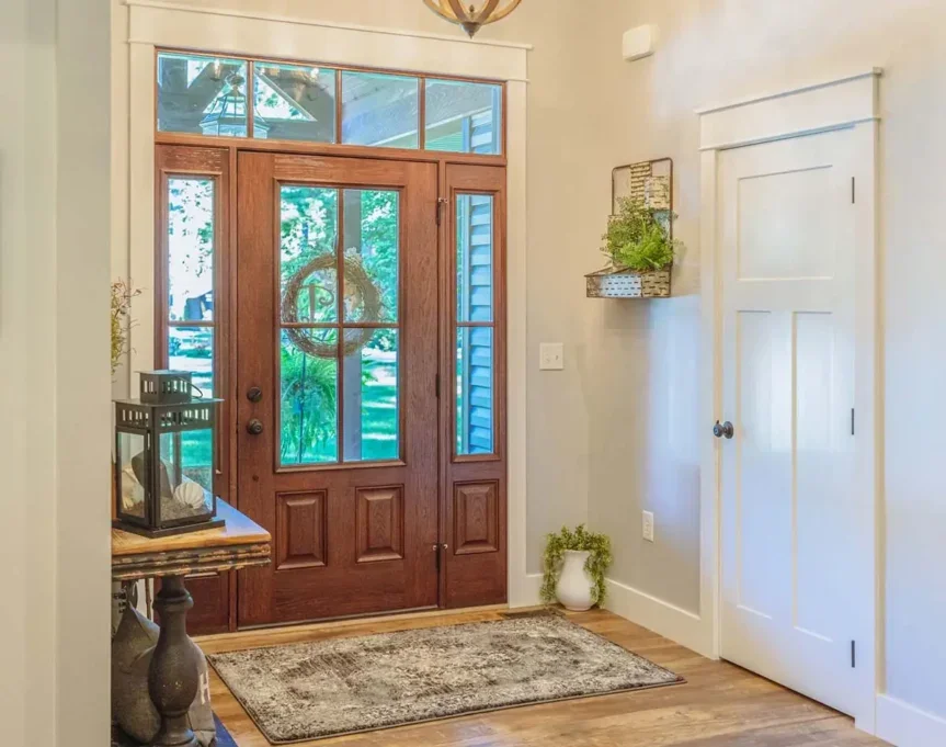 Front door with a window mail slot, decorative wreath, and glass panels, with a small table holding a lantern and decorative item, a potted plant on the floor, a wall-mounted shelf with greenery, and a light-colored door on the right, in a cozy entryway with wooden flooring and a rug.