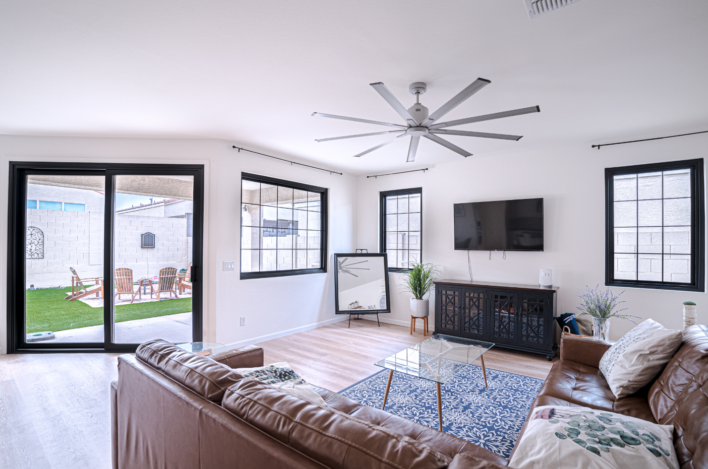 Living room with brown leather sofa, glass coffee table, flat-screen TV on the wall, large windows, potted plants, and sliding door to an outdoor patio with lawn and chairs.
