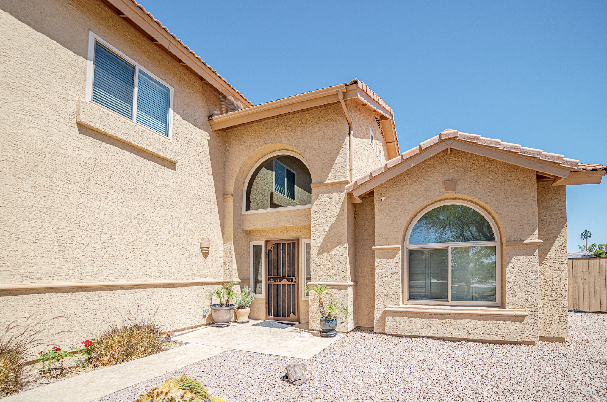 Exterior view of a tan two-story house with a clay tile roof, featuring large arched and rectangular windows, a metal security door, and a gravel yard with potted plants under a clear blue sky.