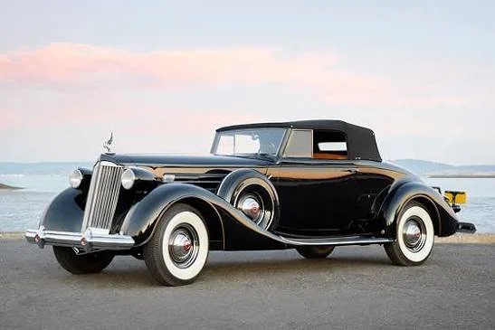 A classic black vintage car parked near water at sunset.