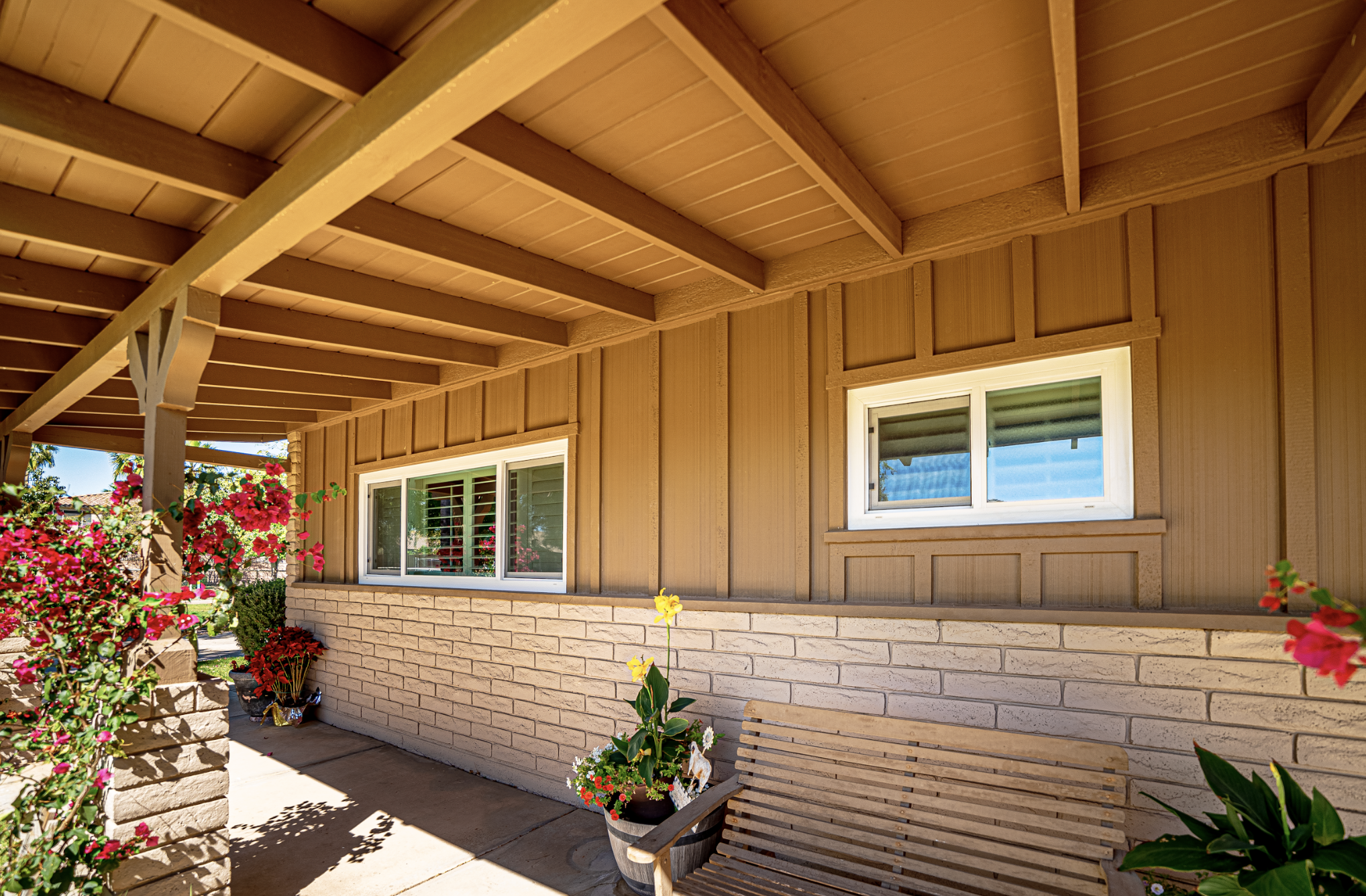 View of a house porch with beige siding, white window frames, potted plants, and a wooden bench, with a flower bush and trees in the background.