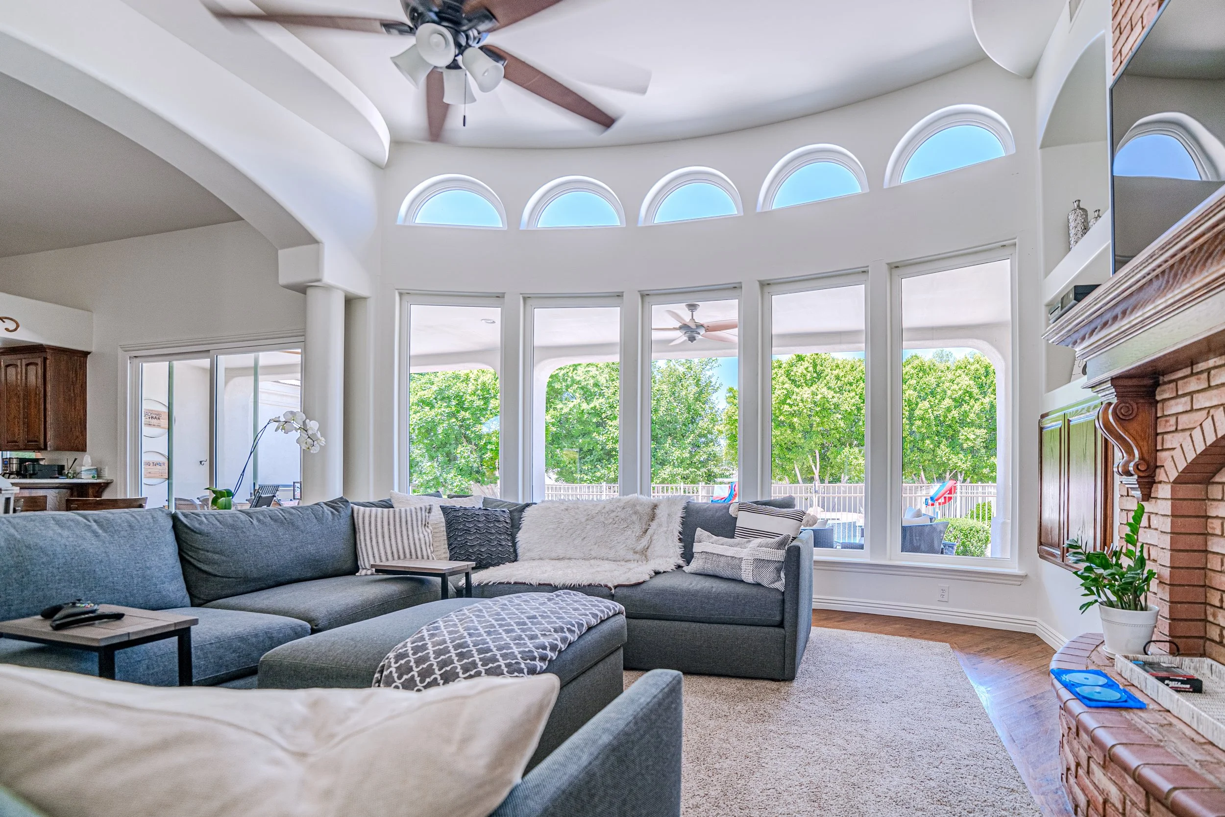 A spacious living room with a large gray sectional sofa, white and patterned throw pillows and a cozy blanket. The room features tall, curved windows letting in natural light, a brick fireplace with decorative items on the mantle, and a view of a backyard with trees, outdoor furniture, and colorful chairs. There is a ceiling fan and a modern light fixture.
