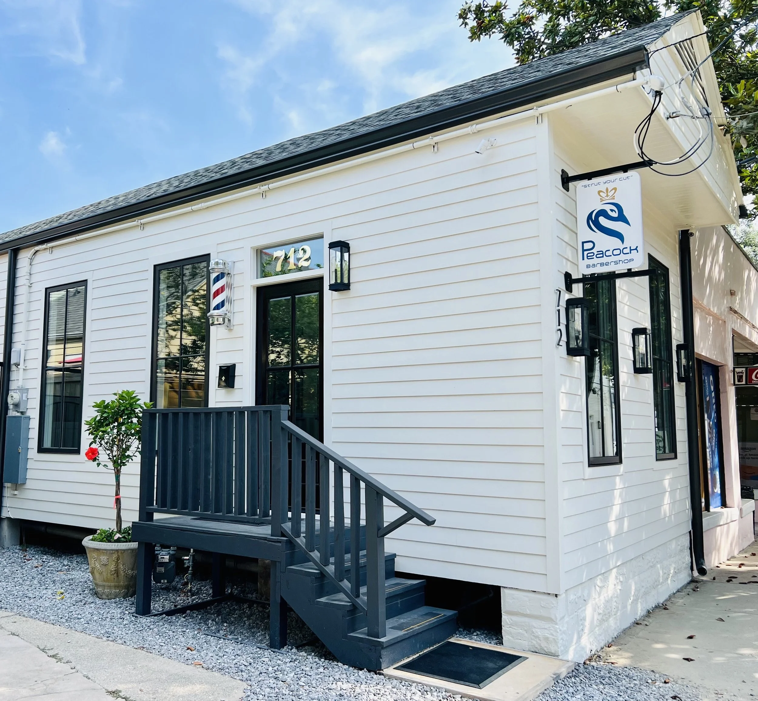 Exterior of a white barbershop with a black door, steps, and a small porch. There is a sign with a stylized swan logo saying 'Peacock Barbershop' and a barber pole next to the door. The building number is 712, and there are two windows with black frames.