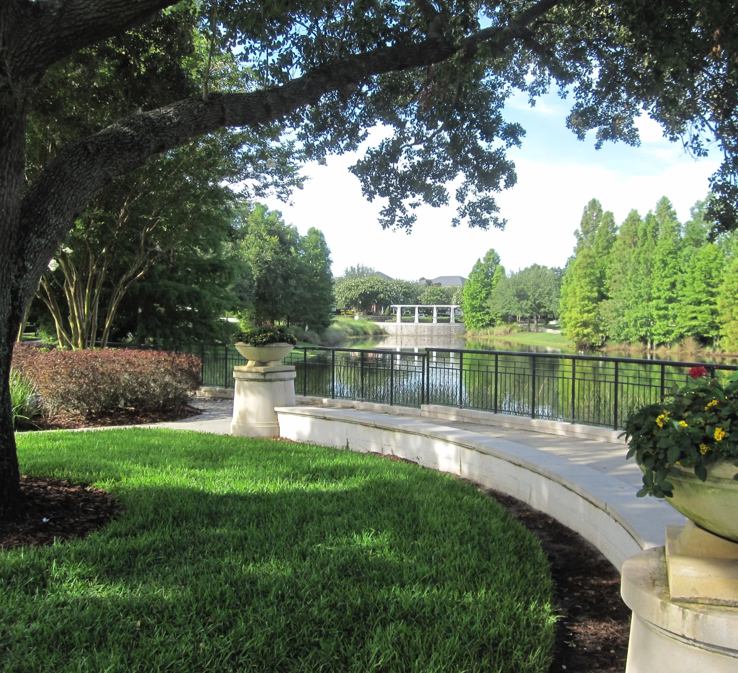 A landscaped garden with a curved paved pathway, lush green grass, potted plants, a large tree providing shade, a calm river with a railing along the edge, and trees on the opposite bank under a partly cloudy sky.