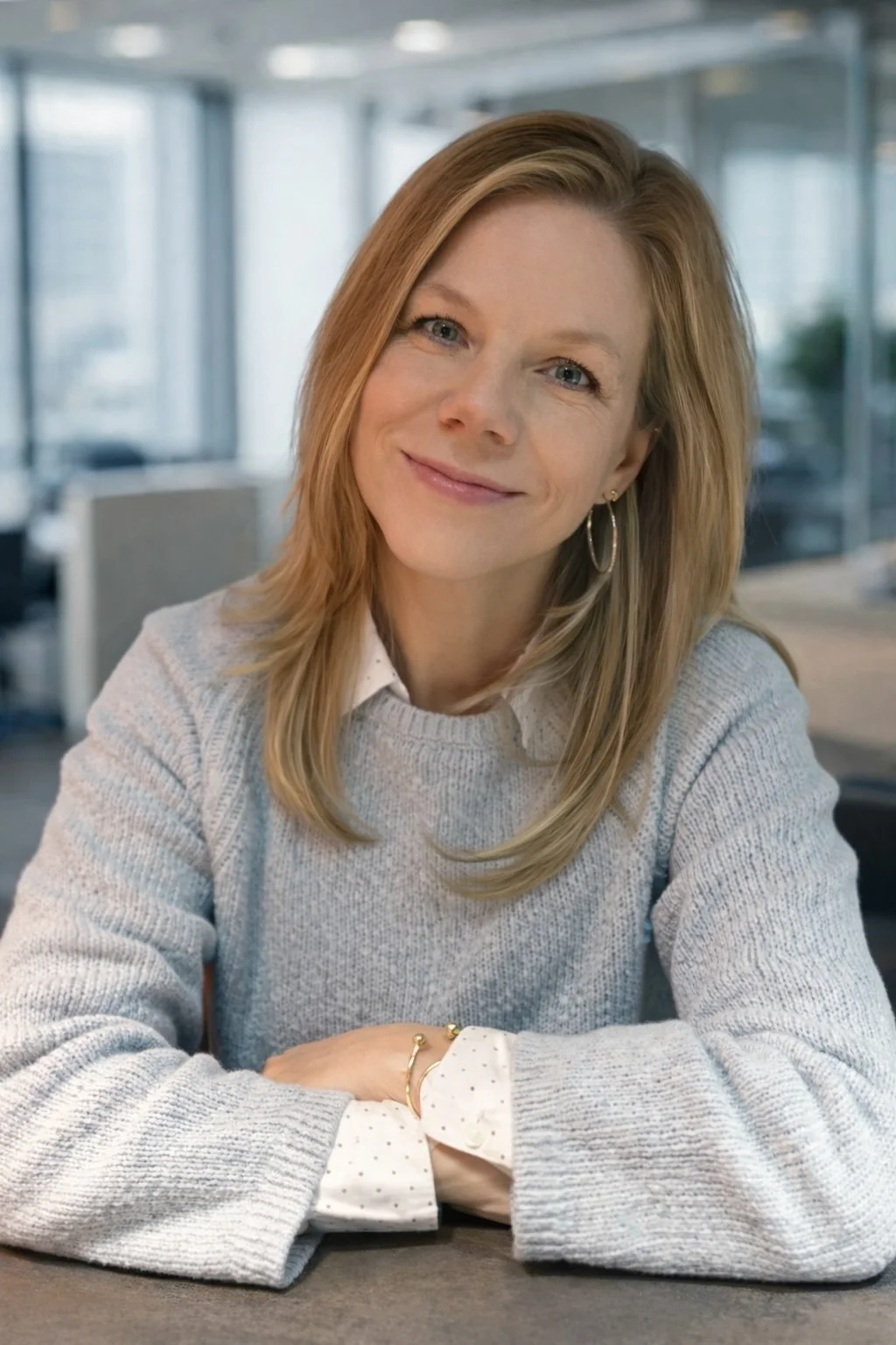 A woman with shoulder-length red hair, wearing a light gray knit sweater over a white shirt with polka dots, smiling with her arms crossed on a table in a modern office setting.