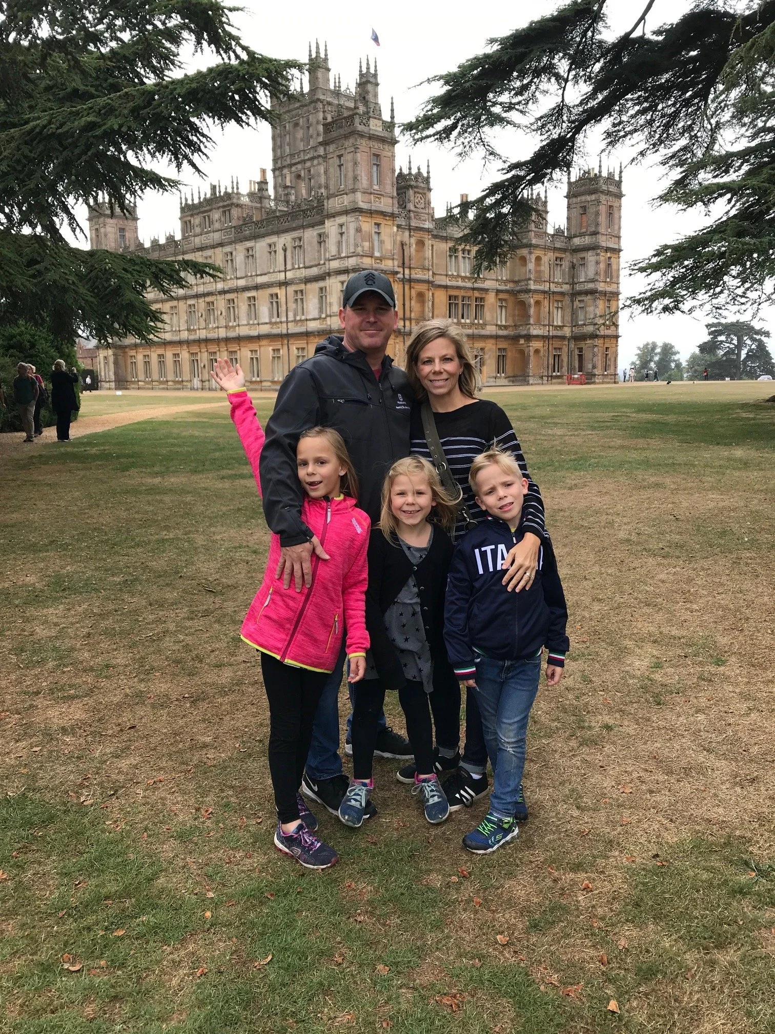 Family of five standing in front of a castle, with two adults and three children, on a cloudy day with trees and grass around.