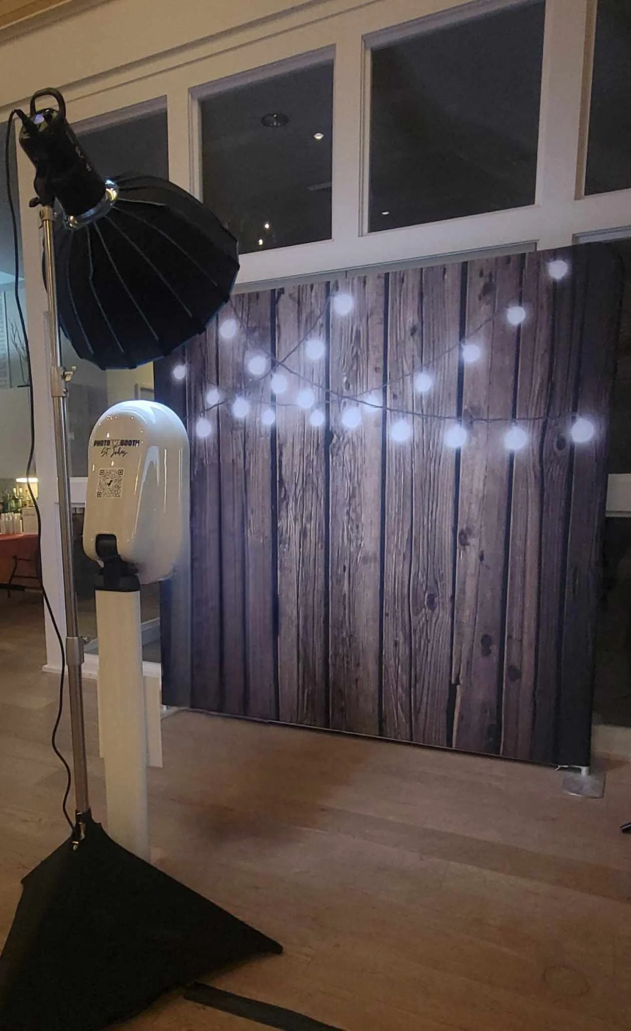 Photo booth setup with wooden backdrop and string lights, a light stand, and a table with props like hats and crowns beside it.