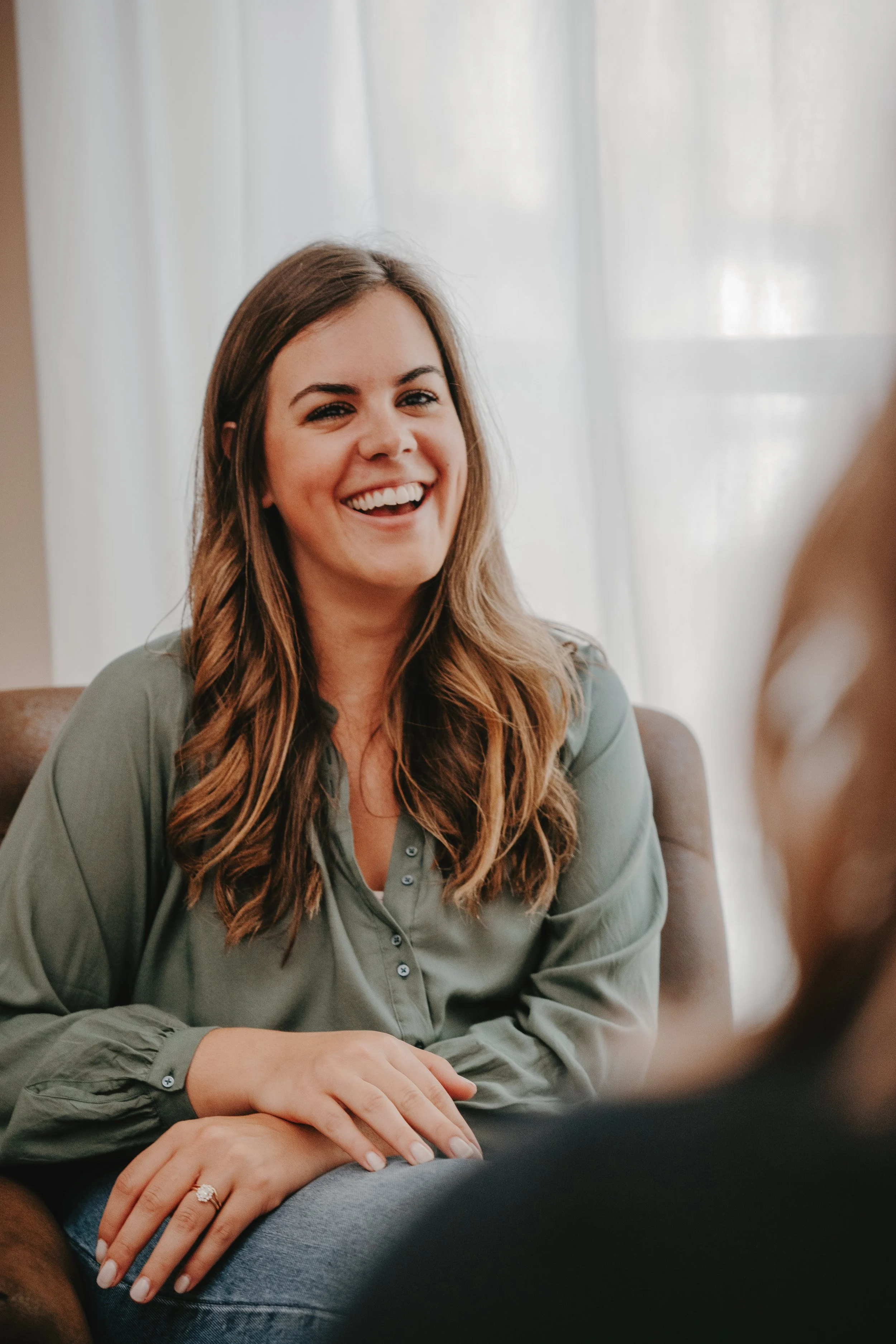 woman with brown curly hair laughing while sitting in a chair talking to a woman in the distance