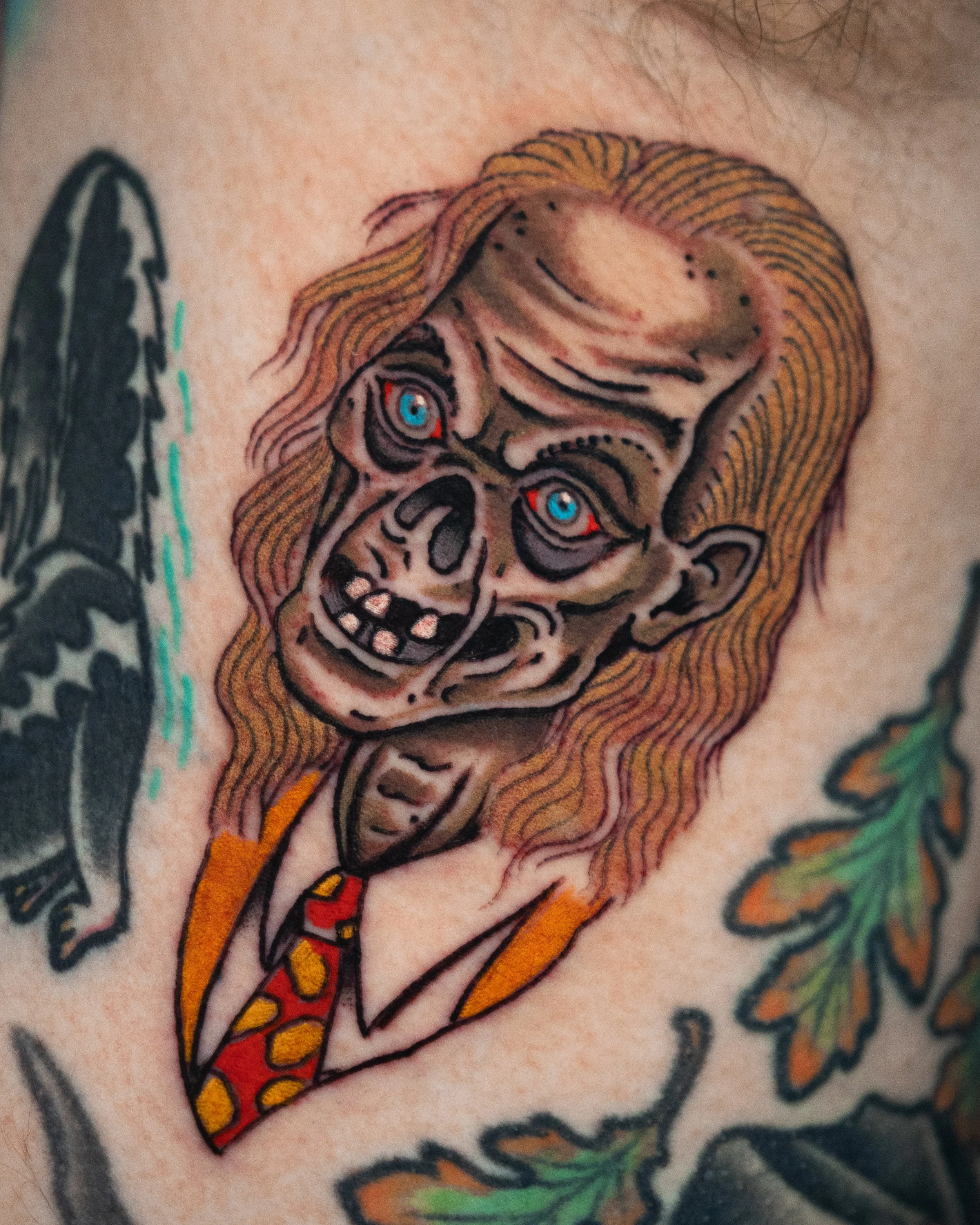 A colorful tattoo of a zombie-like face with blue eyes, open mouth, and red tongue, surrounded by leaves.