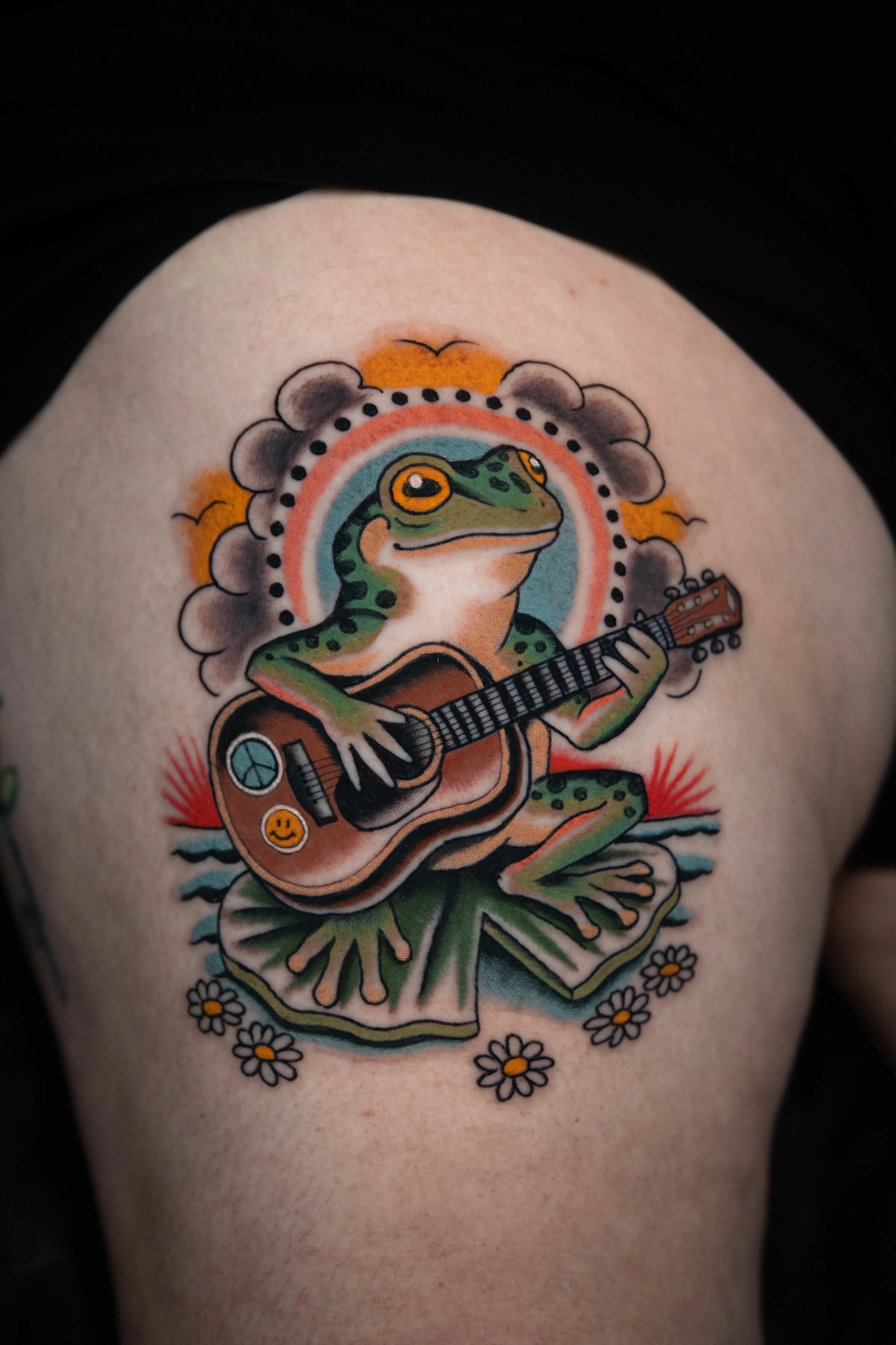 Tattoo of a frog playing an acoustic guitar, sitting on lily pads with daisies, sunset, and clouds in the background. Done at San Luis Tattoo Company by Danny Derrick in San Luis Obispo, CA SLO.