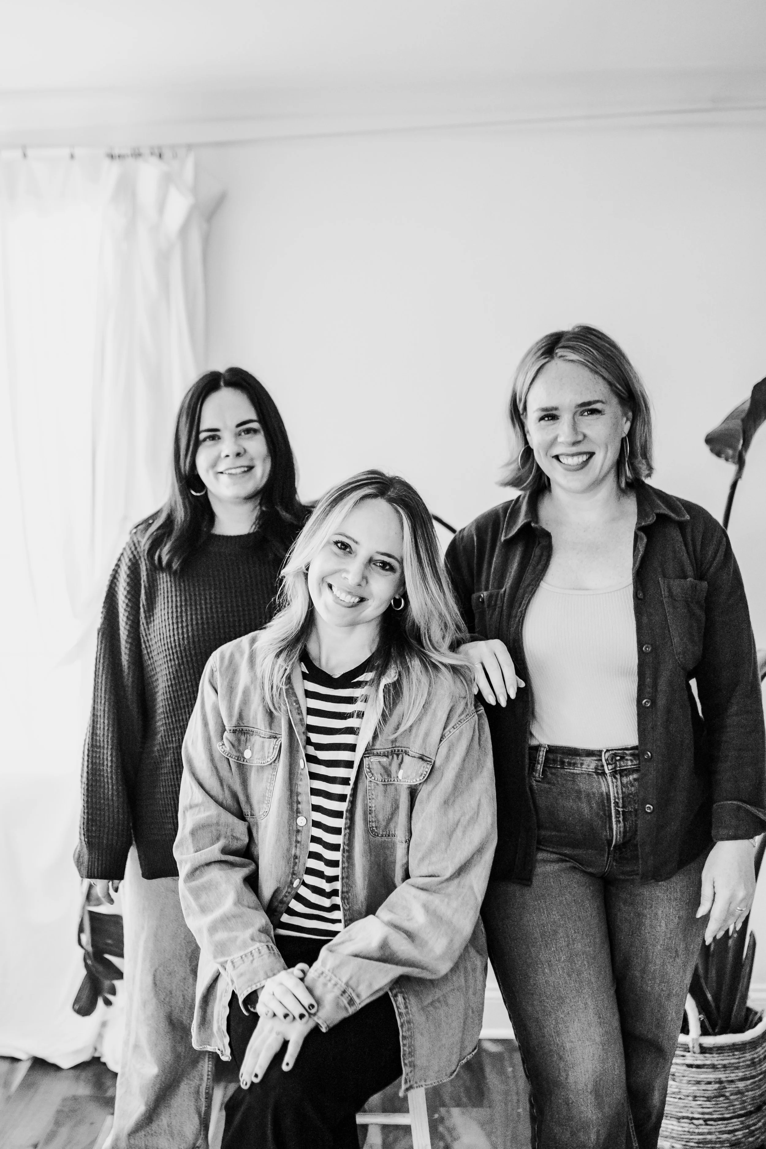 Three women smiling, posing together indoors. The woman in front is seated and making a peace sign, wearing a striped shirt and a denim jacket. The other two women are standing behind her, one on the left with curly hair in a sweater, and one on the right with short hair in a button-up shirt.