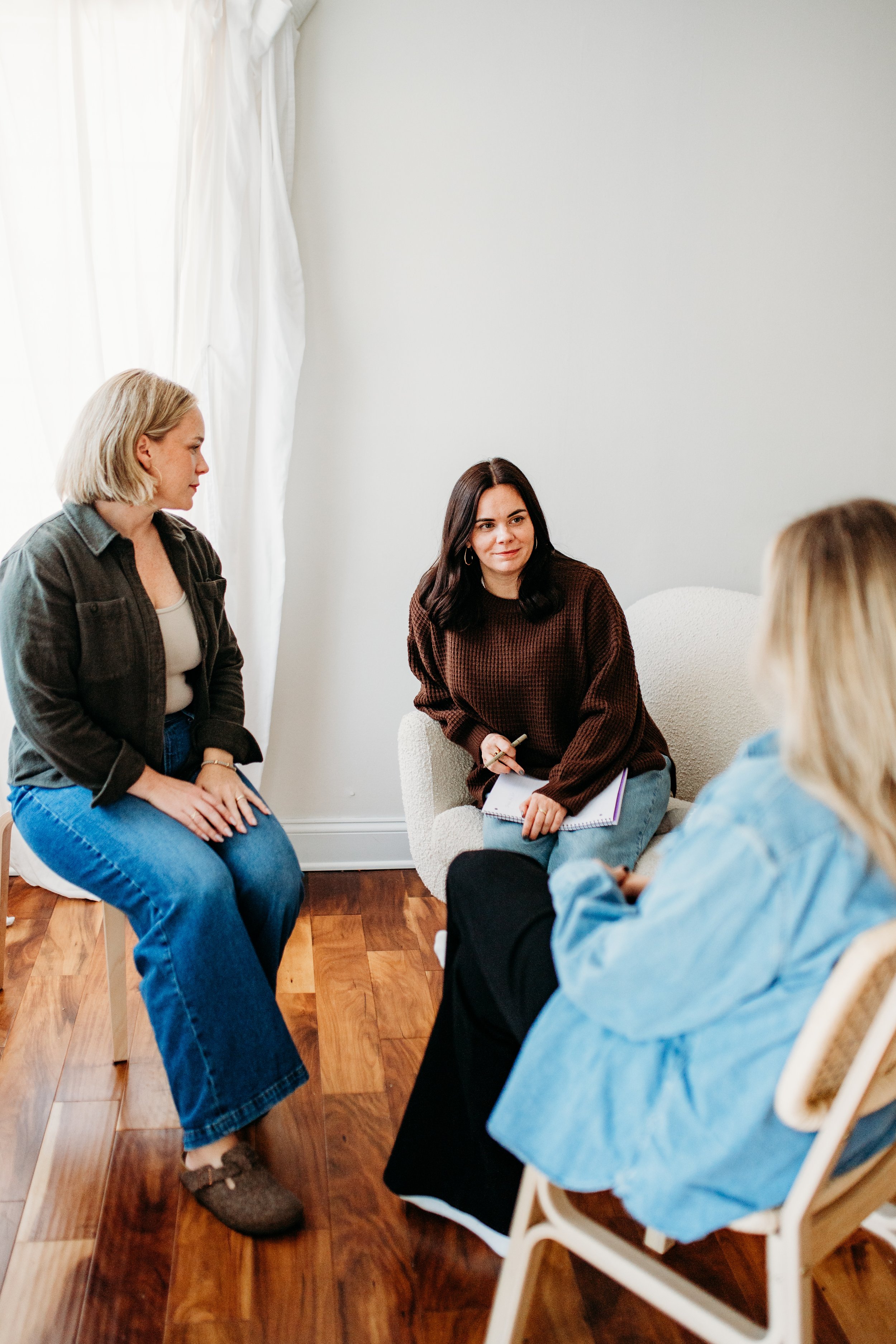 Three women having a discussion in a bright room with white walls and hardwood floors.