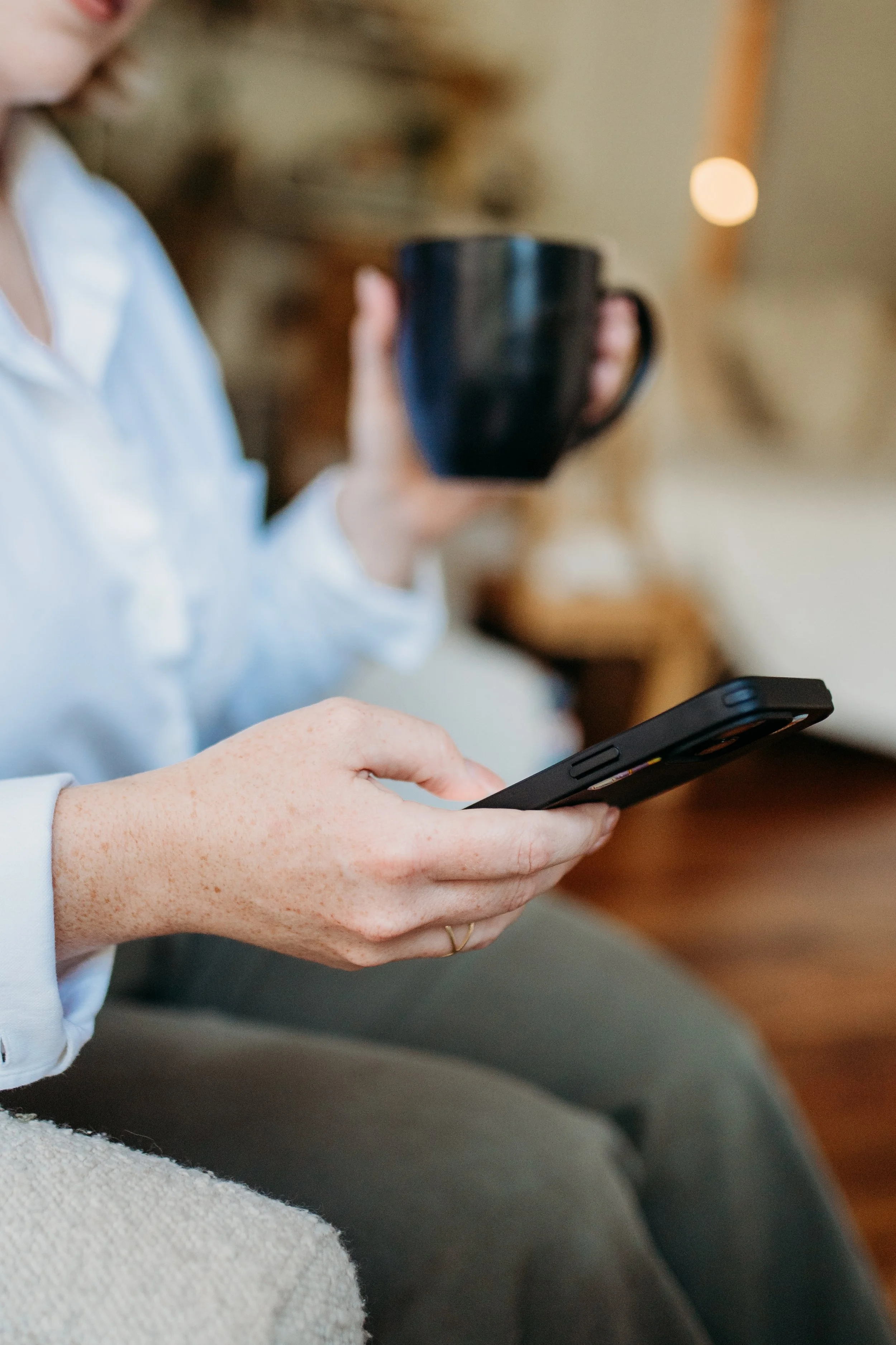 A person sitting on a couch holding a smartphone in one hand and a black mug in the other, with a blurred background indoor setting.