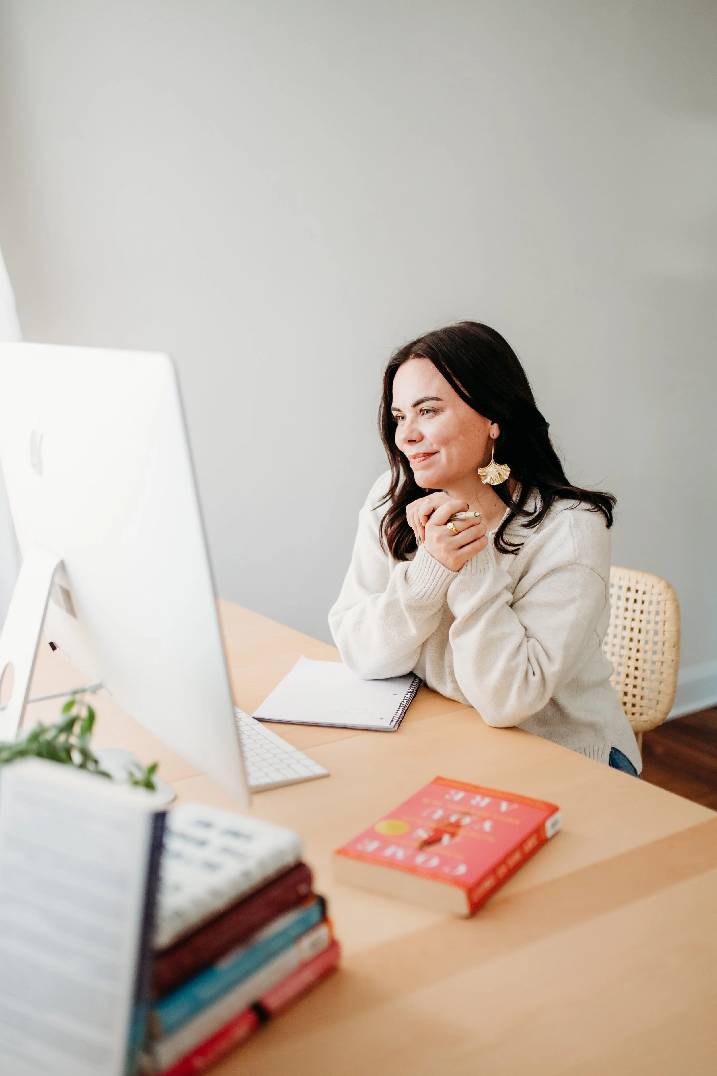 A woman sitting at a desk, working on a computer with a notepad, books, and a book on the desk, in a bright room.