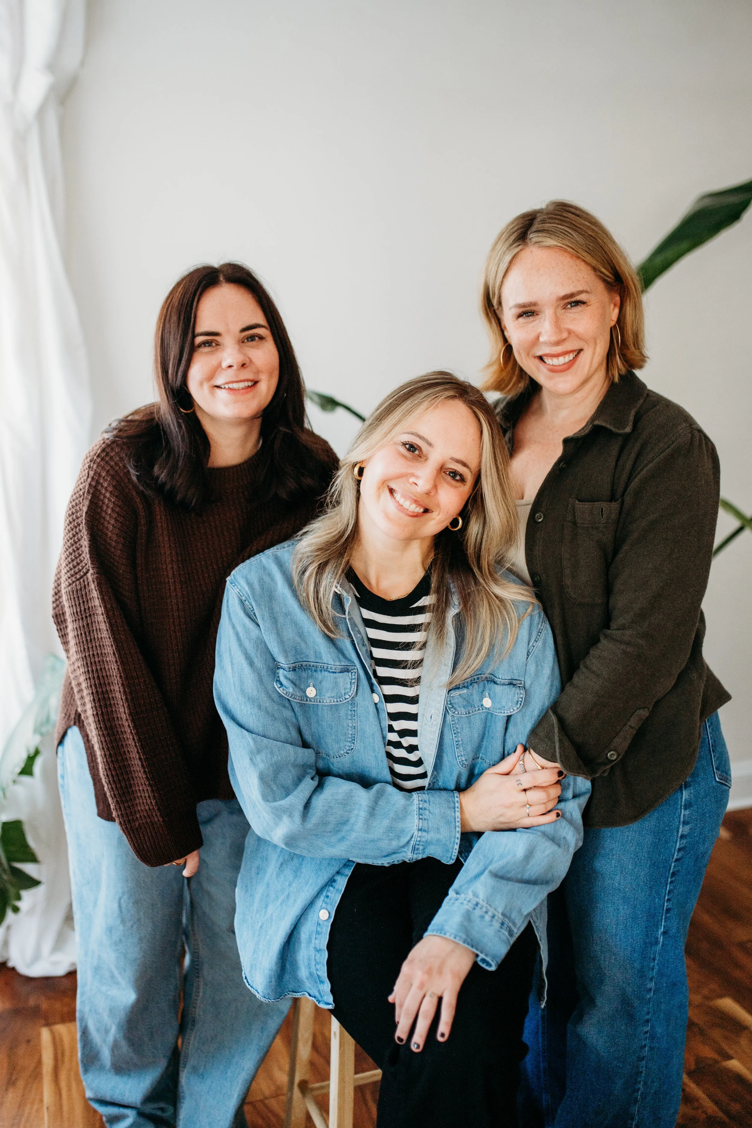 Three women smiling, one sitting in front and two standing behind in a well-lit room with plants and wooden floor.