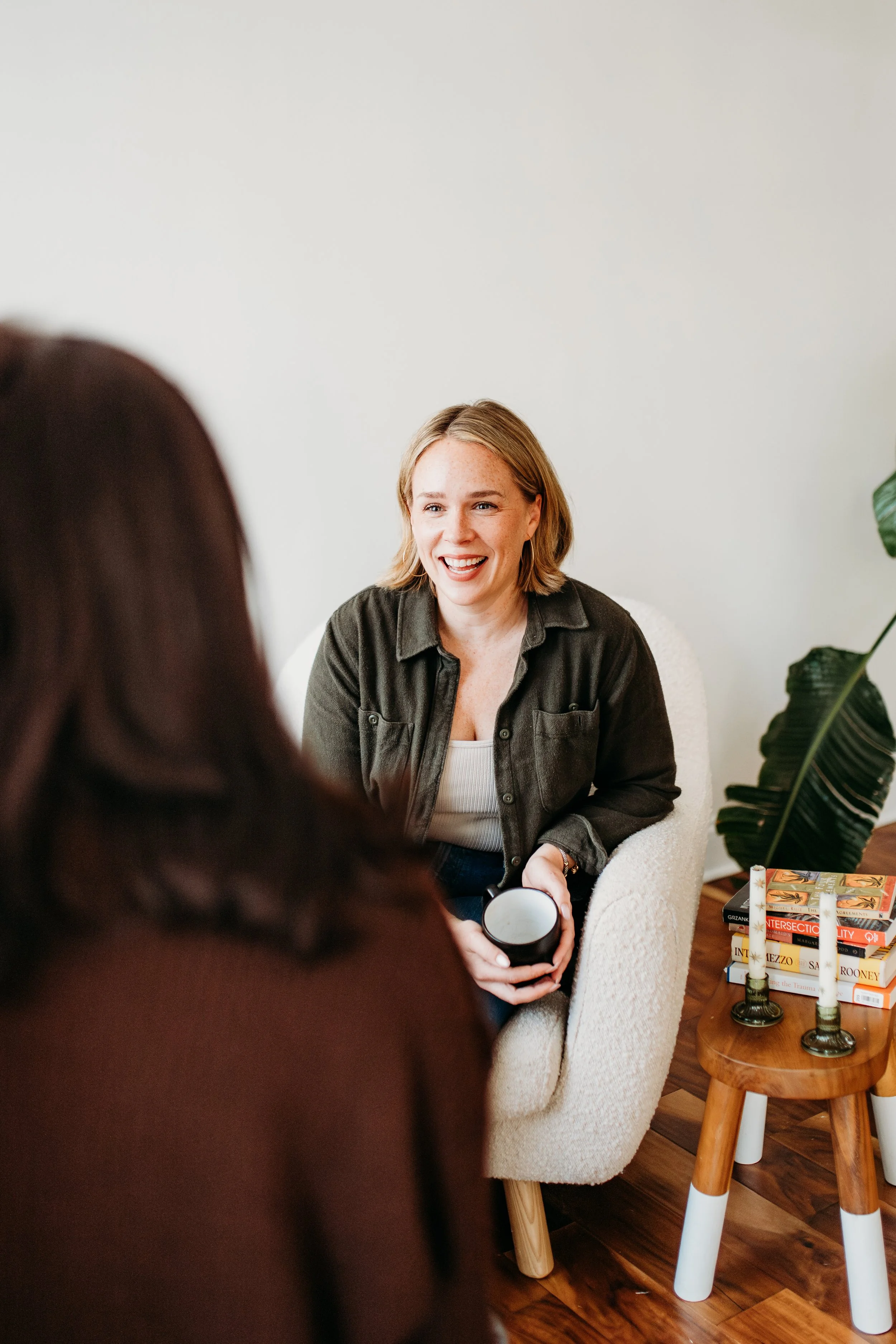 Two women having a conversation in a cozy room, one woman smiling and holding a coffee mug, the other person with long dark hair visible from the back.