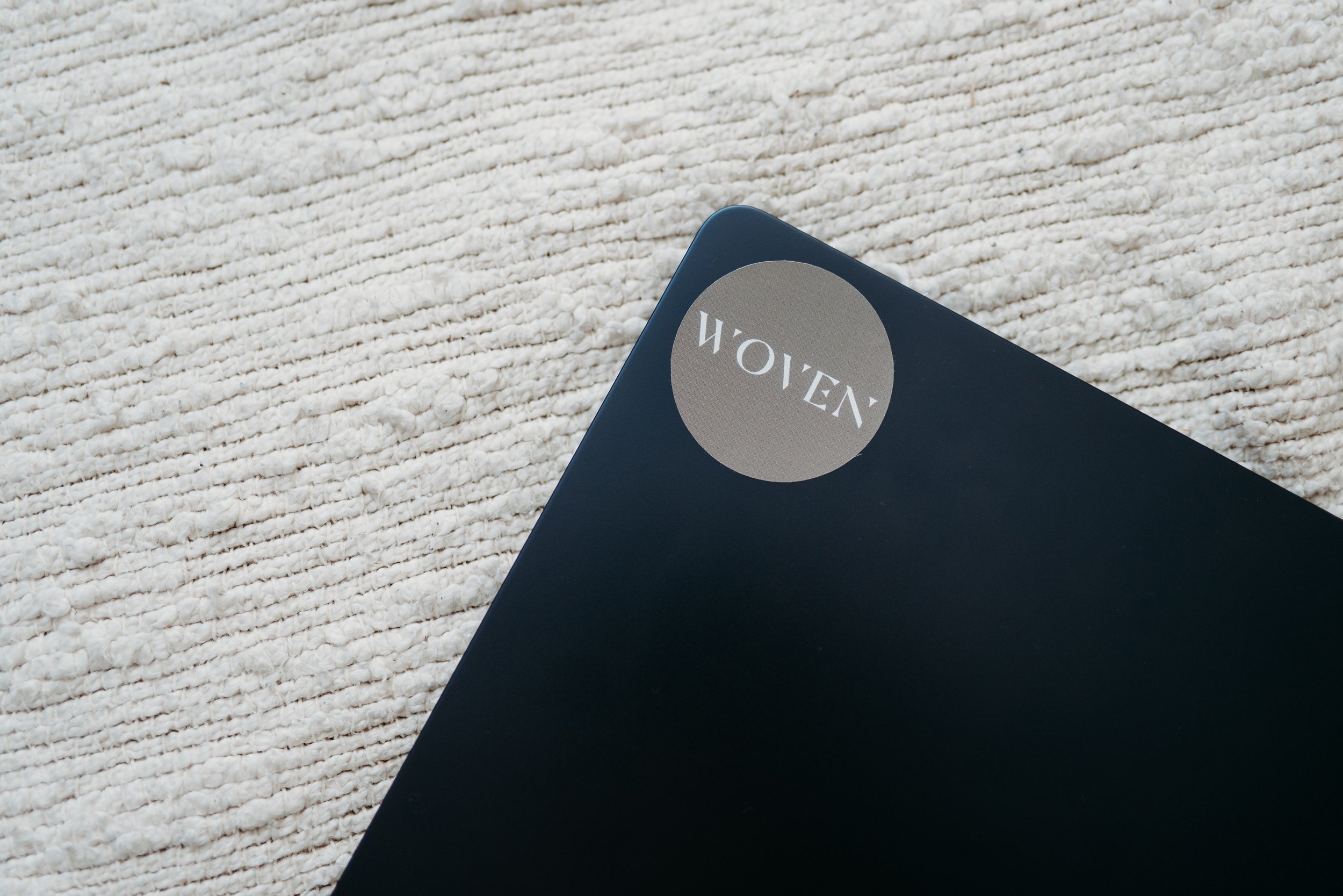 Close-up of a black notebook with a gray circular sticker that says 'HOVEN' on a textured white surface.