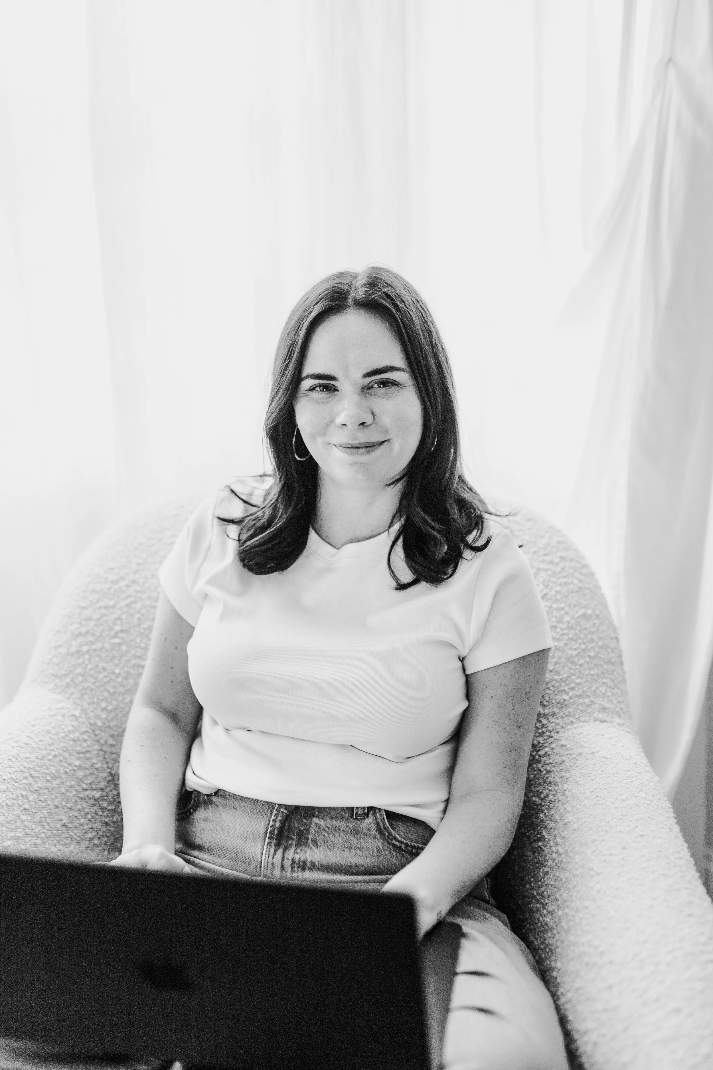Black and white photo of a woman with shoulder-length dark hair sitting on a textured armchair with a laptop in front of her, in a well-lit room with curtains in the background.