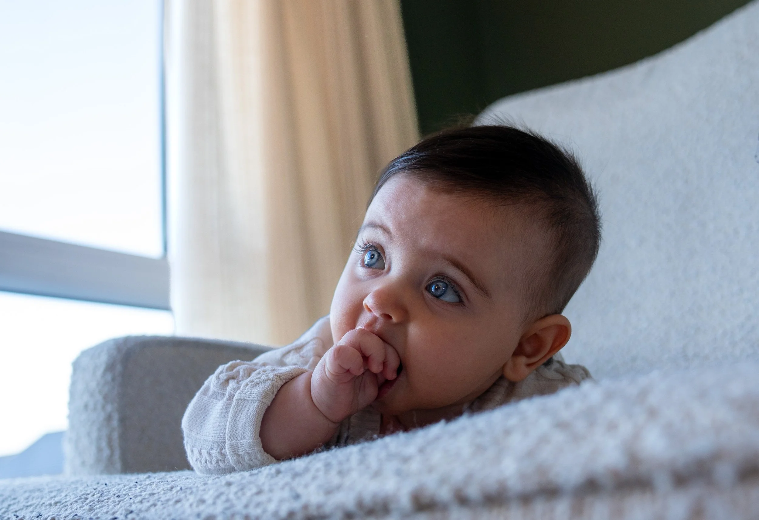Close-up of a baby with blue eyes lying on a light-colored blanket, sucking on fingers in a cozy indoor setting with a window in the background.