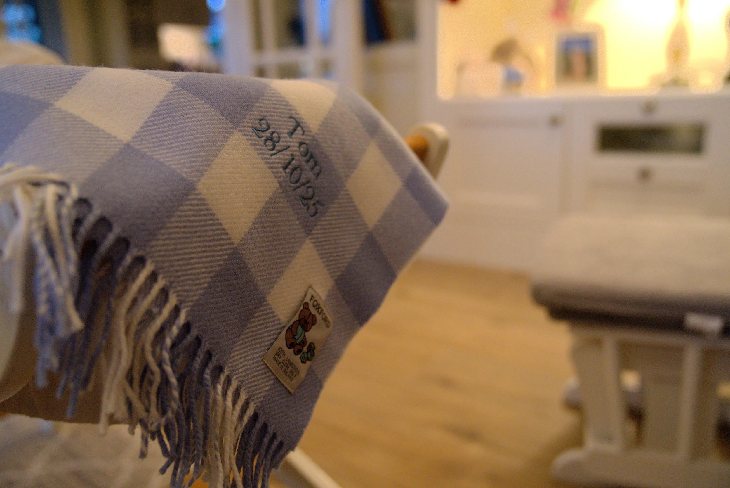 Close-up of a blanket with a blue and white checkered pattern and fringe, with a label showing "25/189" and a cartoon fox character, draped over a wooden chair in a cozy room.
