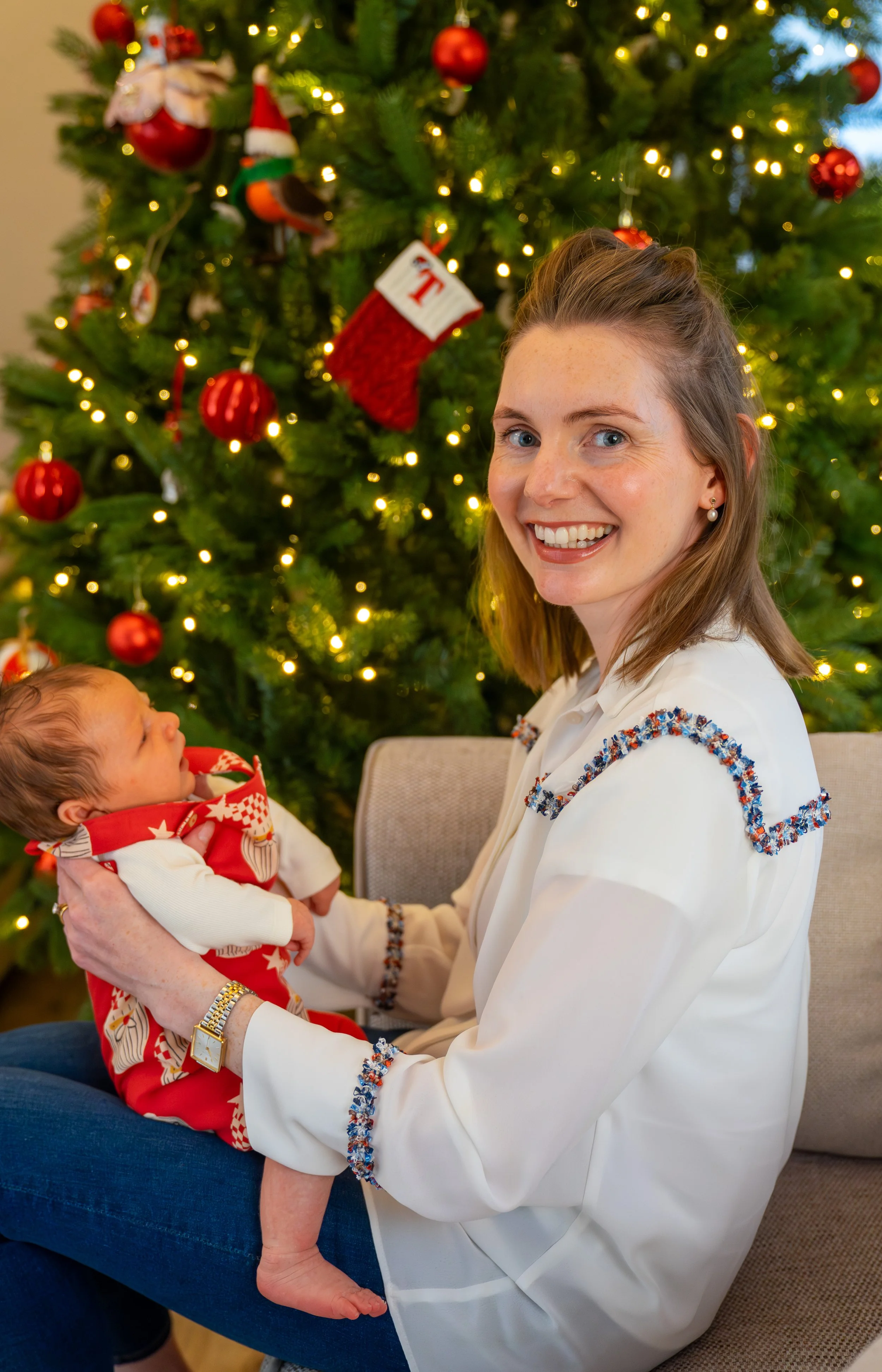 A smiling woman holding a baby in front of a decorated Christmas tree with red ornaments and lights.