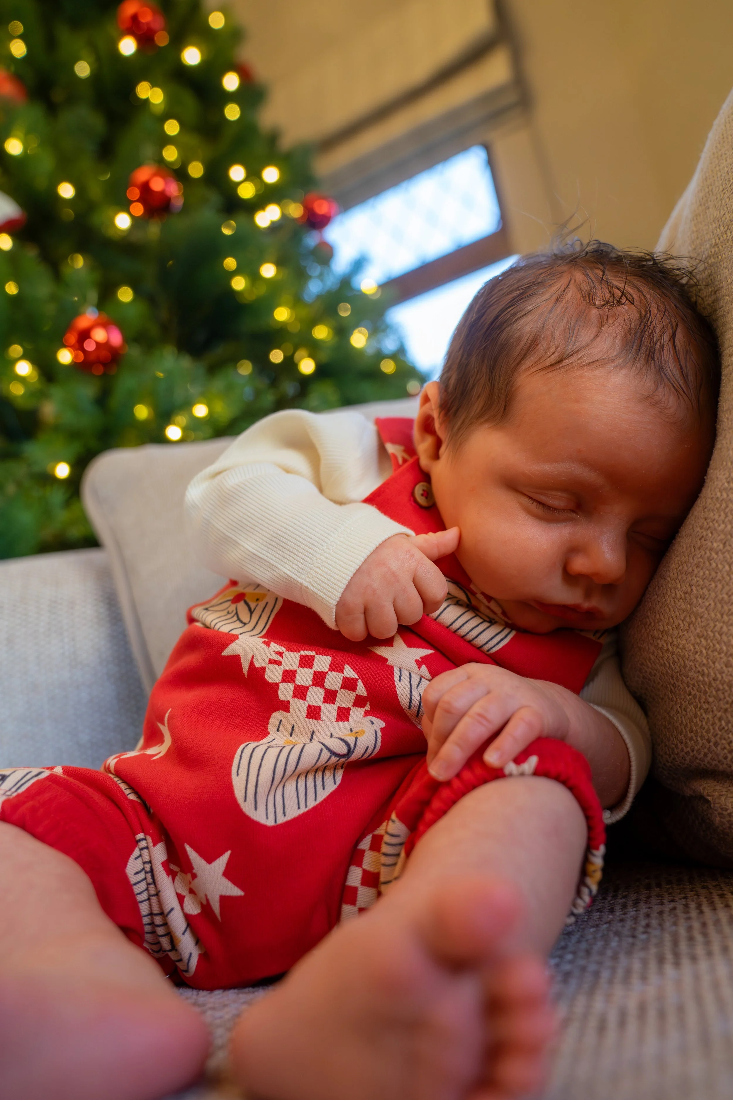 A sleeping baby lying on a couch next to a Christmas tree decorated with lights and red ornaments.