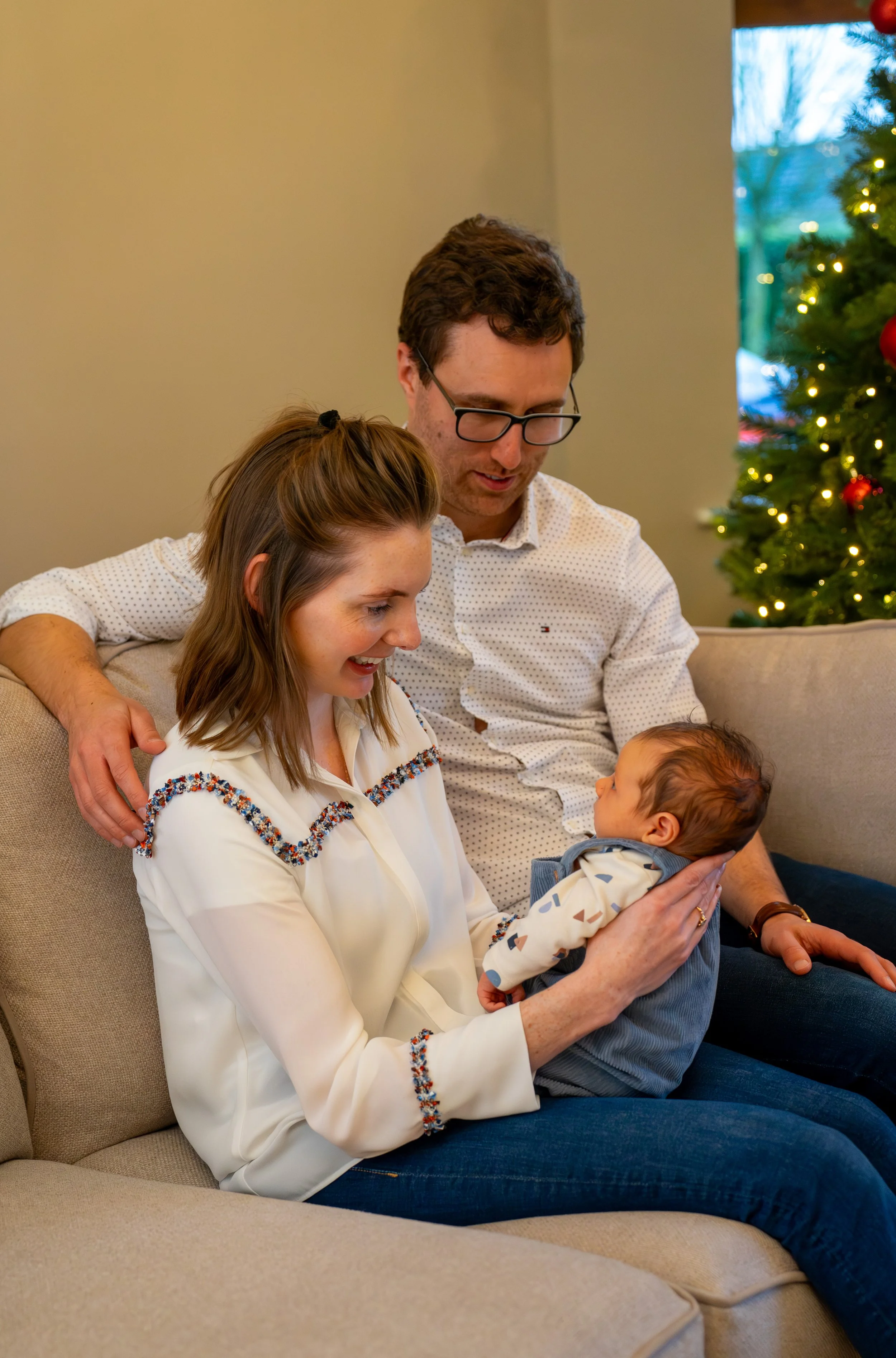 A family of three, including a woman, a man, and a baby, sitting on a beige sofa near a decorated Christmas tree with Christmas lights in a living room.