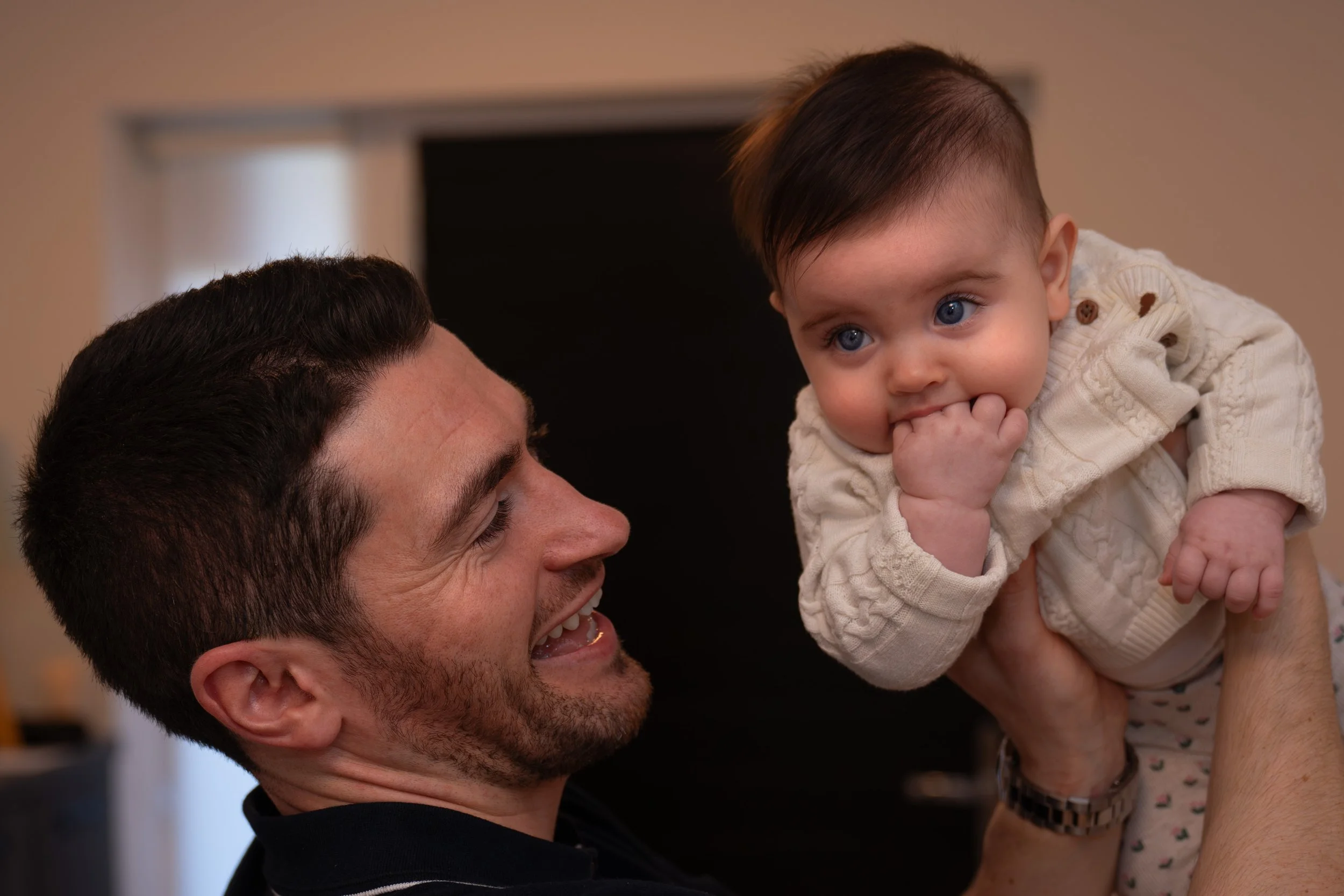 A man is smiling and holding a baby girl with blue eyes and dark hair.