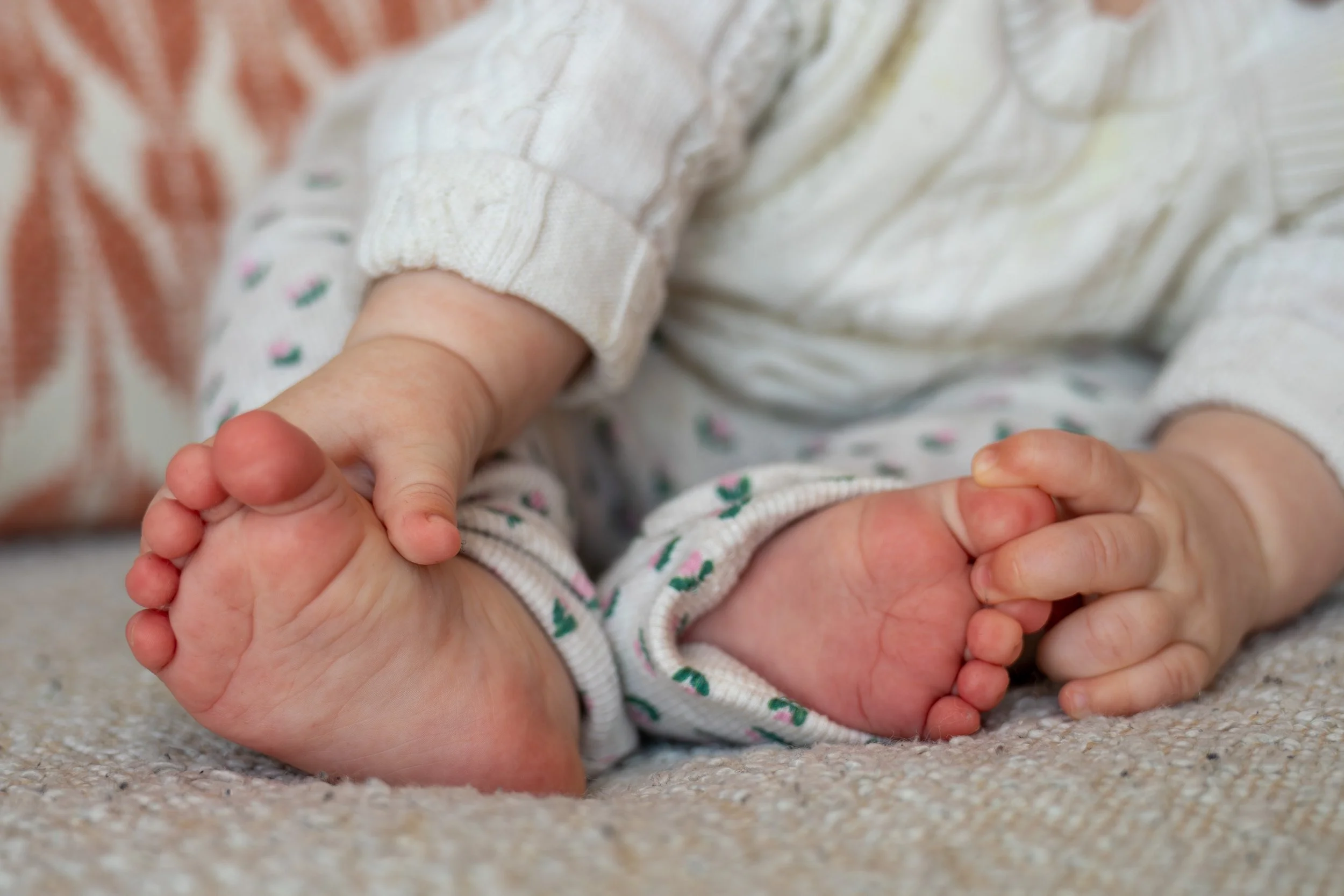 Close-up of a baby's feet resting on a textured beige surface, with the baby's hands gently holding one foot. The baby is wearing white socks with small pink and green floral patterns, and a light-colored, long-sleeved shirt.