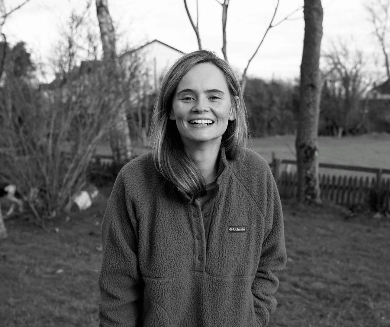 Black and white photo of a smiling woman with shoulder-length hair wearing a Columbia fleece jacket, standing outdoors with trees and a fence in the background.