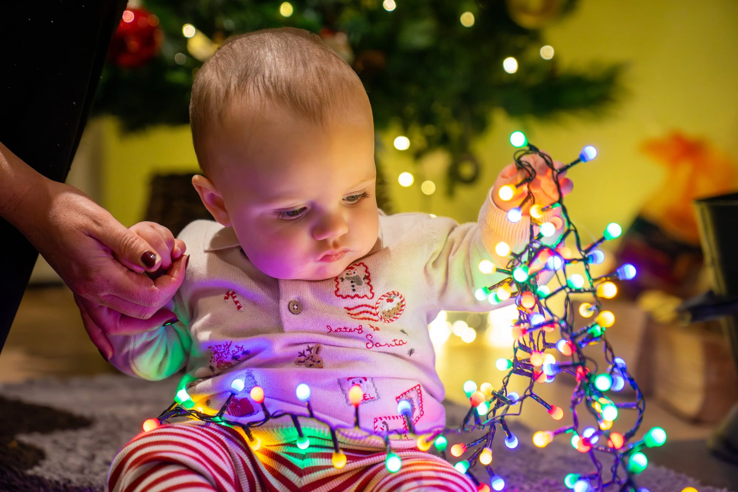 A young child sitting on the floor, holding and examining colorful Christmas lights, with a decorated Christmas tree in the background.