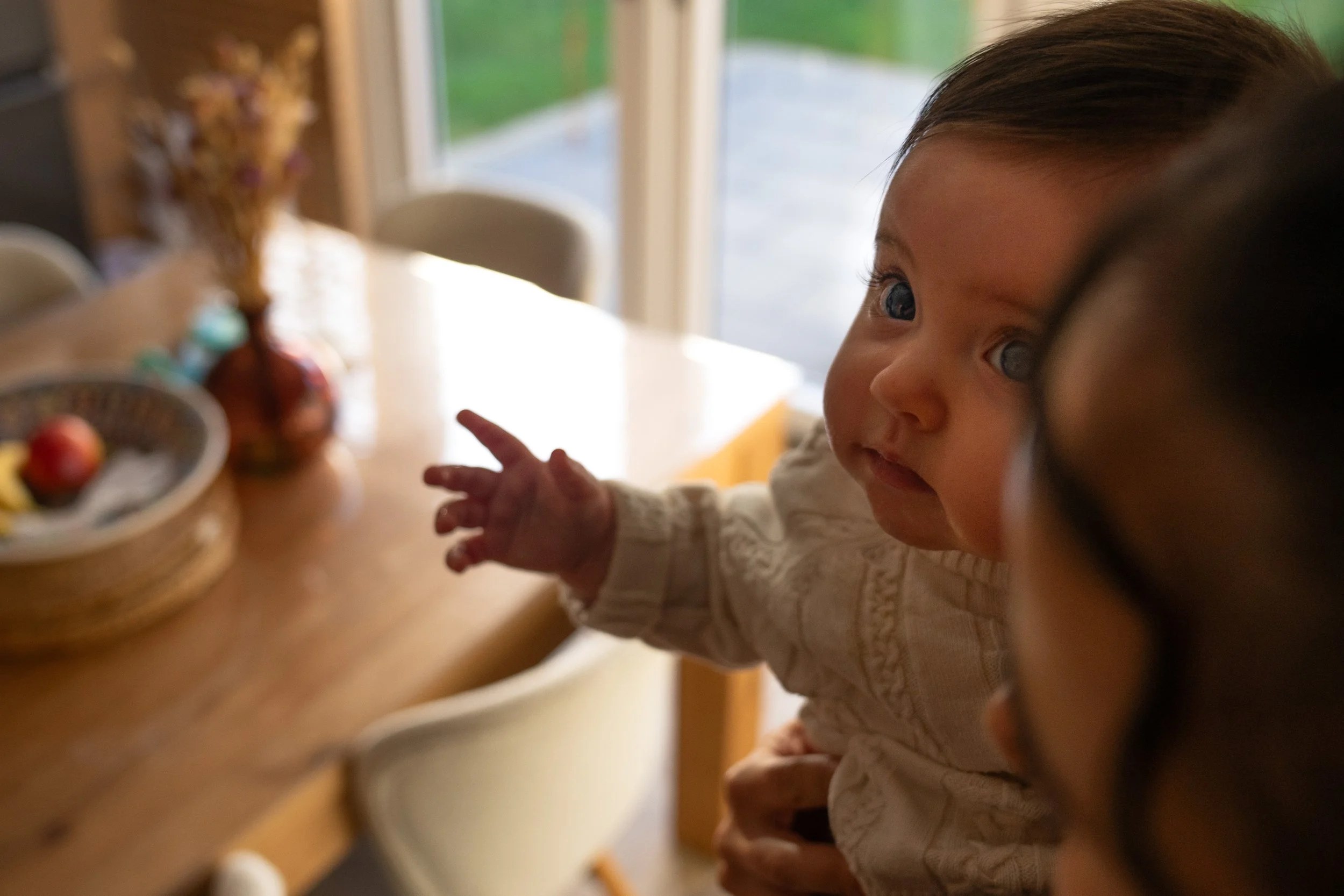 A baby with blue eyes wearing a cream-colored sweater pointing at a flower arrangement on a wooden table, with a woman partially visible nearby.