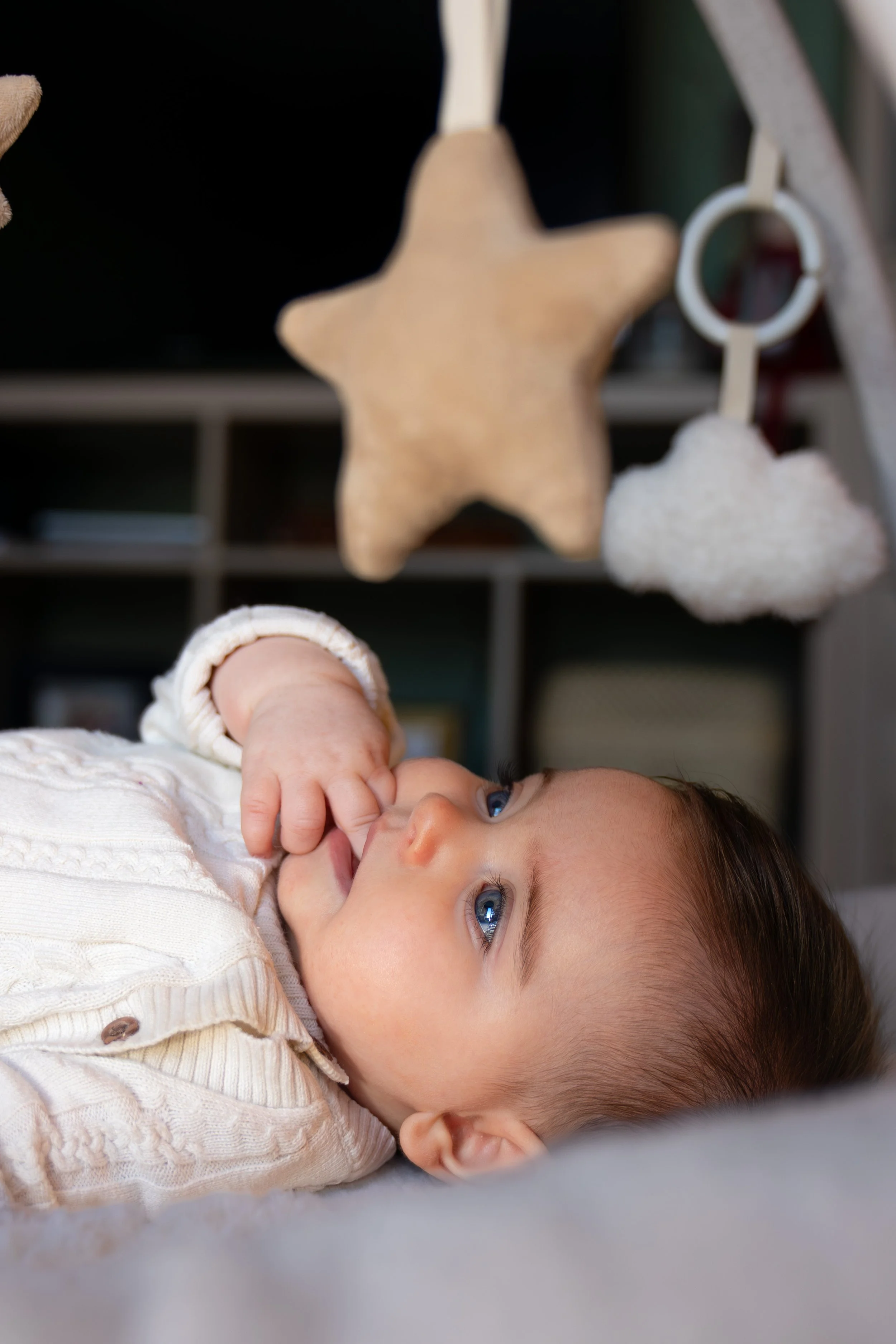 A young child with blue eyes lying on a bed, looking at hanging plush toys, including a star-shaped toy, in a cozy nursery.