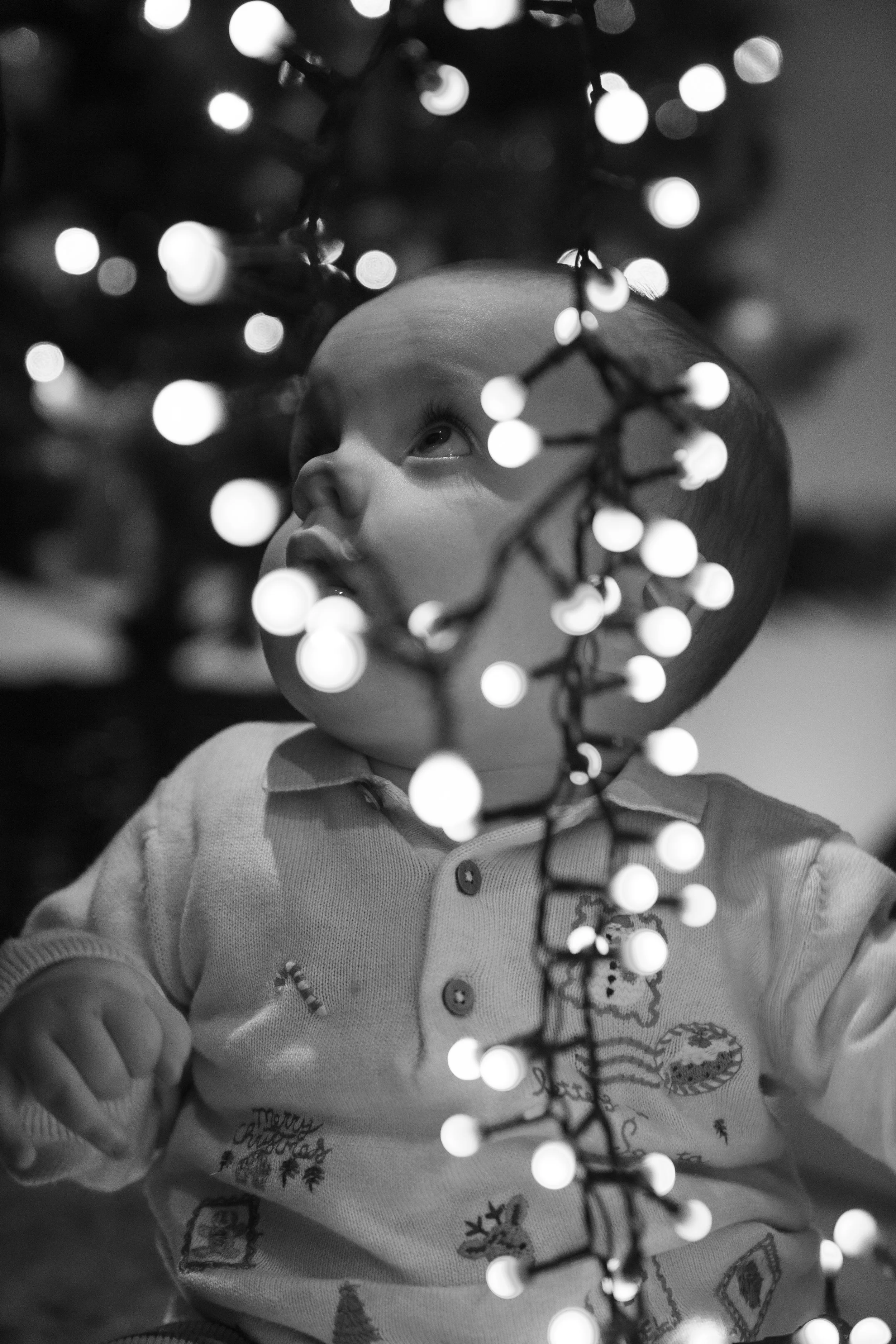 Black and white photo of a young child looking up through Christmas tree lights.