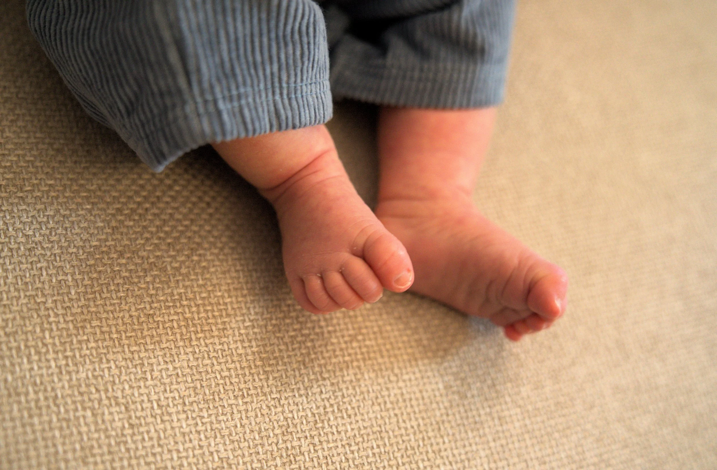 Close-up of a baby's legs and feet resting on a beige textured surface, with the baby wearing gray striped pants.