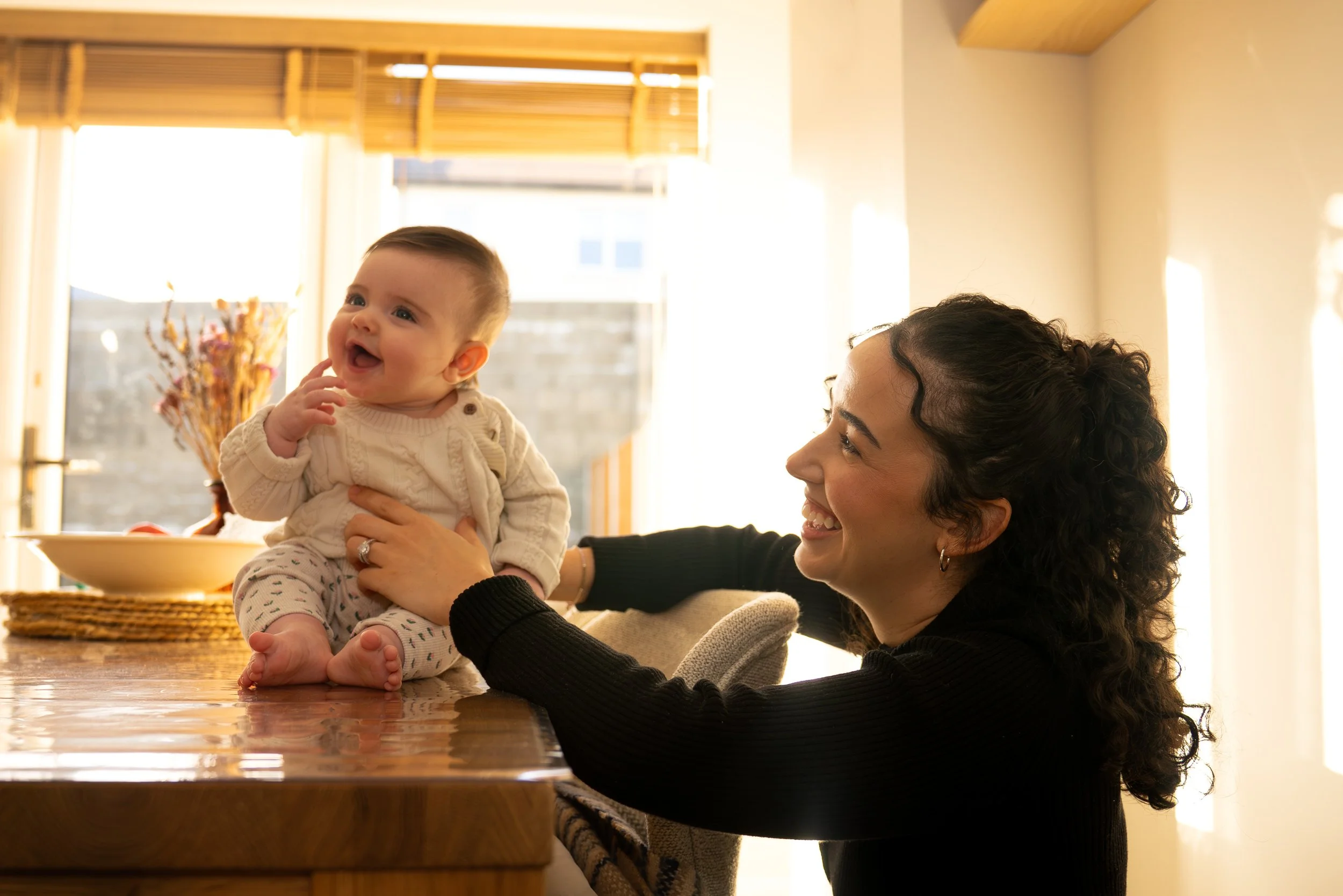 A woman with curly hair smiling at a baby sitting on a table in a sunlit kitchen.
