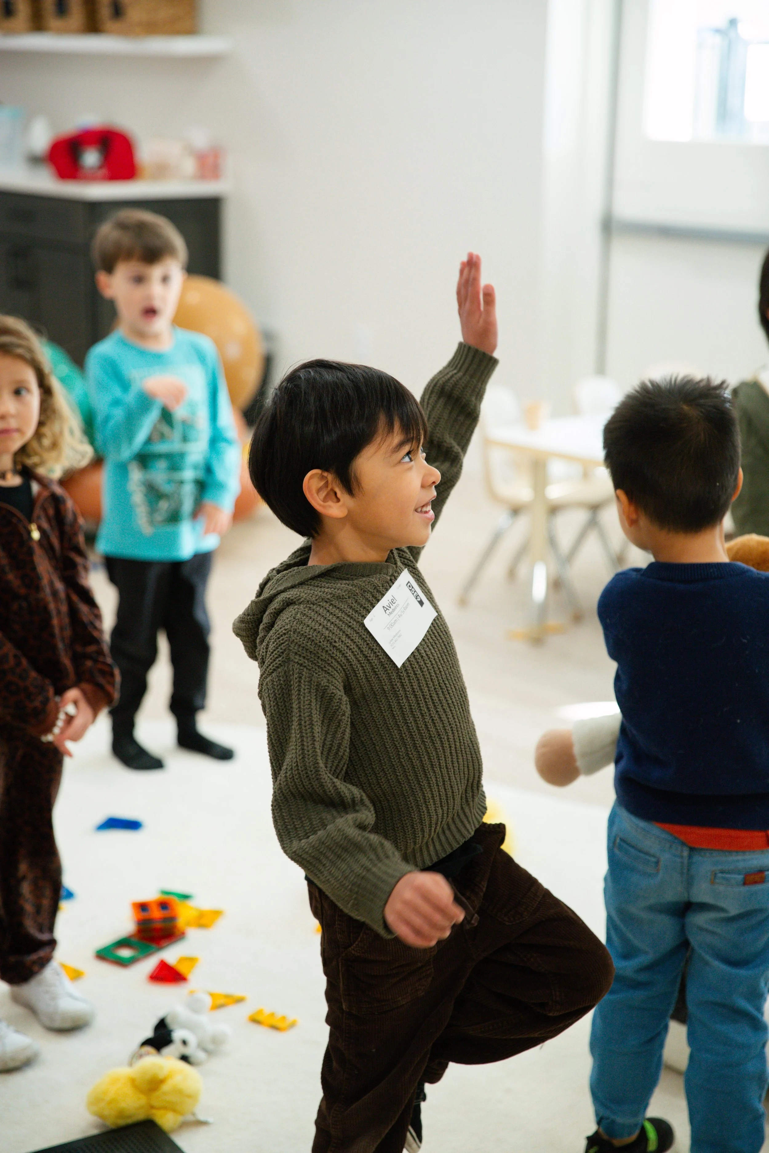 Young boy playing with toys on a blue carpet in a playroom with other children in the background.
