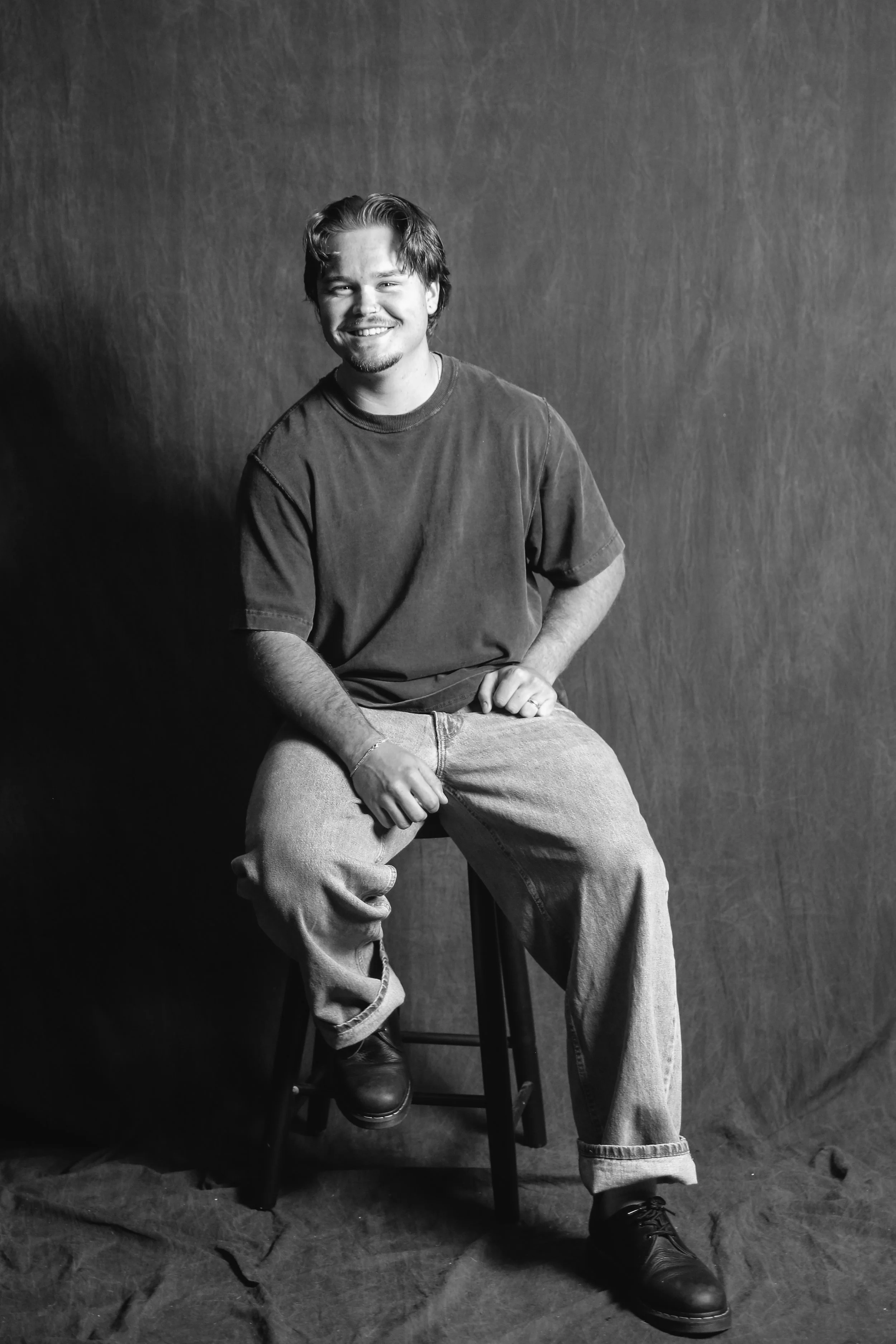 A black-and-white portrait of a young man with a slicked-back hairstyle, beard, and mustache, wearing a dark knit sweater and light-colored pants, sitting on a stool against a dark background.