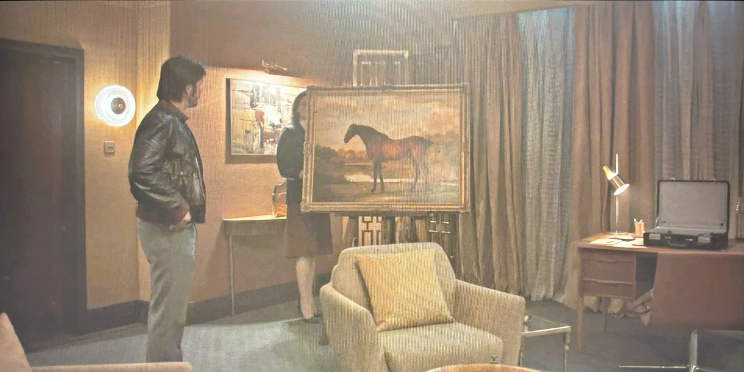 A man in a leather jacket and beige pants stands in a living room, looking at a woman viewing a painting of a horse on an easel. The room has warm lighting, a beige armchair with a yellow pillow, a desk with a turntable, and curtains covering the window.