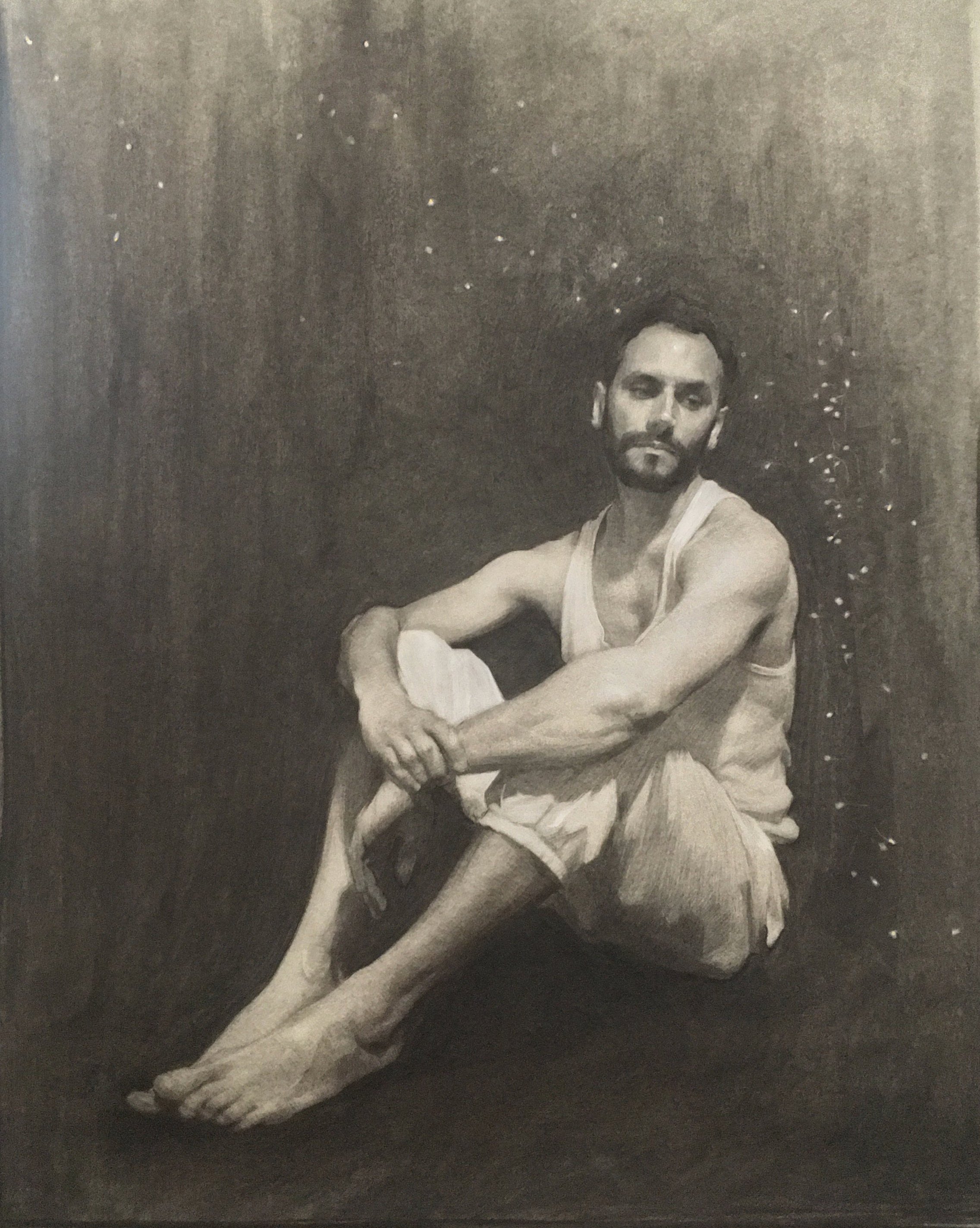 A black-and-white portrait of a man with a beard, sitting barefoot on the floor with his knees bent and arms resting on them. He is wearing a sleeveless shirt and shorts, gazing thoughtfully into the distance against a dark, textured background.