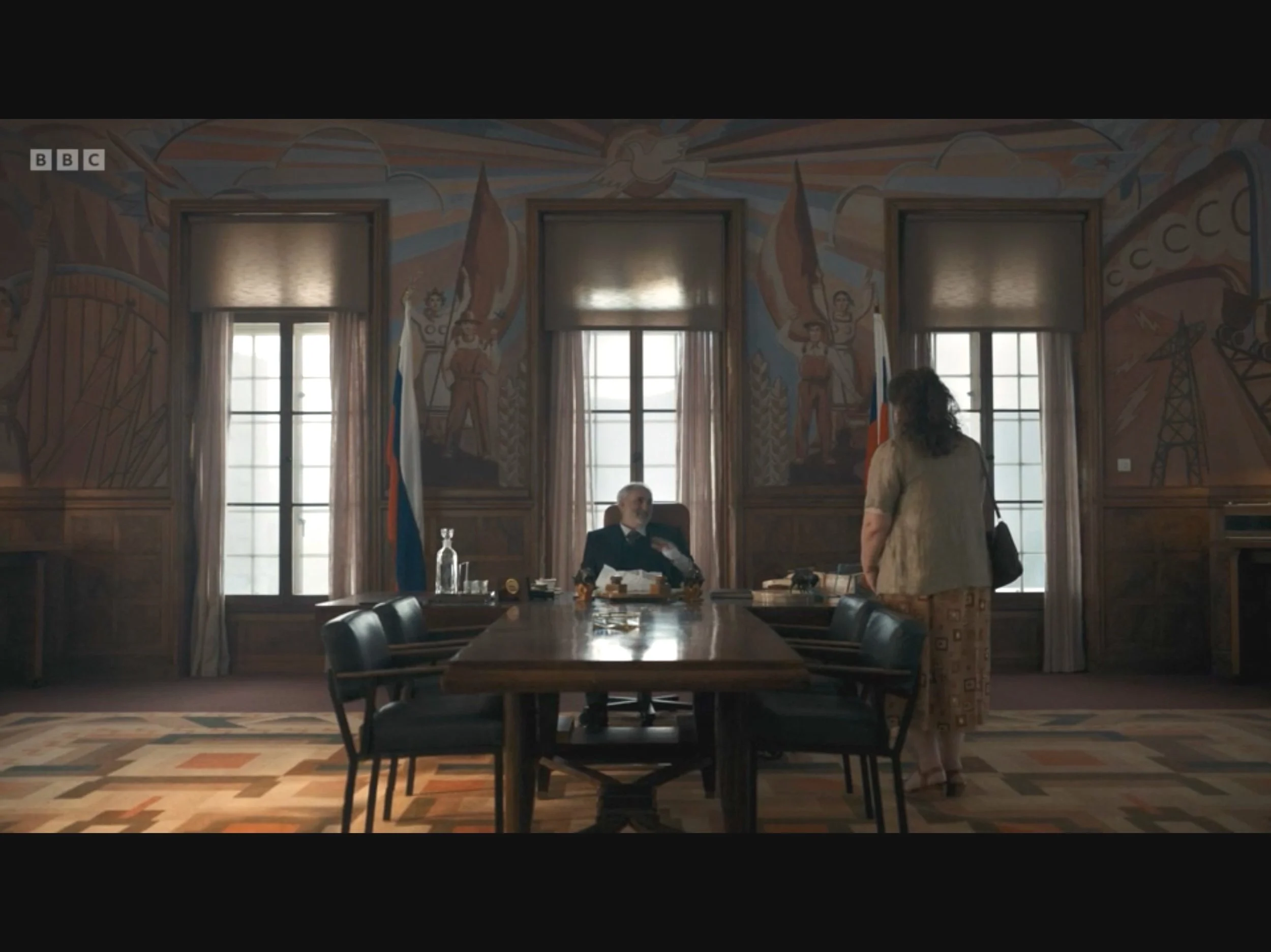 A man sitting at a large wooden conference table in an ornately decorated room with murals of workers and industrial imagery on the walls, and a woman standing and talking to him.